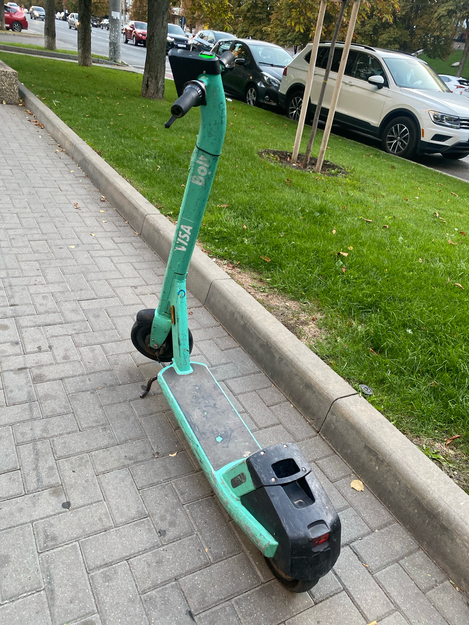 An abandoned green electric scooter with "Bolt" and "VISA" branding is parked on a sidewalk of interlocking gray pavers. The scooter has a black platform, black handlebars with a brake lever, and a black rear casing with a red reflector. To the right of the sidewalk is a grassy area with fallen leaves and trees, bordering a street with several cars parked and driving. The scene appears to be in an urban setting in Chișinău, Moldova, given the visual cues of the architecture, vegetation, and vehicles.