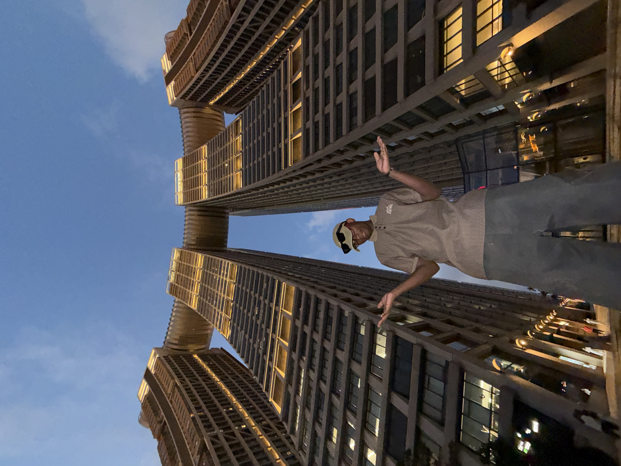 In the Yuzhong District of China, a person stands on steps in front of two towering, modern buildings connected by a skybridge. The person, wearing a light brown collared shirt, dark pants, sunglasses, and a cap, faces the camera with arms outstretched and palms up. The sky is a gradient of deep blue transitioning to lighter hues near the horizon, with scattered clouds. The buildings are illuminated, casting a warm glow on their facades and windows. The perspective is from a low angle, emphasizing the grandeur of the architecture.