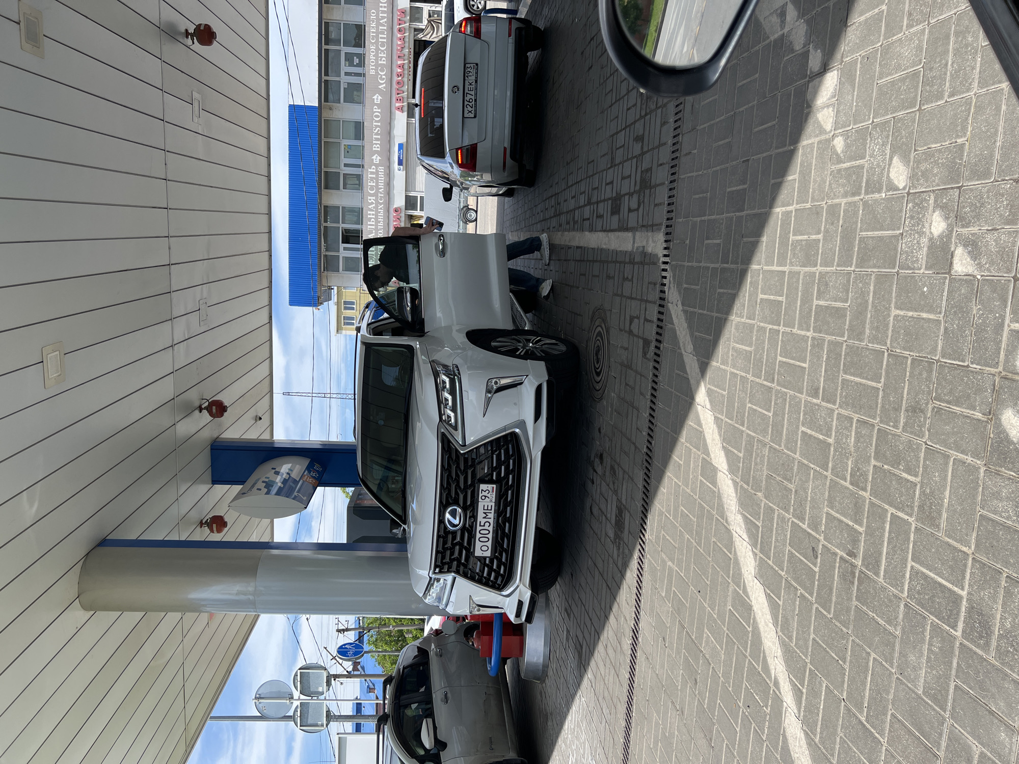 This media file captures a sunny daytime scene at a gas station in Krasnodar, Russia, viewed from inside a vehicle.
In the foreground, a white Lexus SUV, featuring a distinctive large grille, is positioned at a fuel pump. Its Russian license plate reads "O005ME 93". A person, possibly the driver, is visible standing near the open driver's side door of the Lexus, suggesting they are in the process of refueling the vehicle.
Behind the Lexus, a white Skoda Octavia is also present, displaying the Russian license plate "X267EK 193". The cars are parked on a paved surface made of interlocking grey pavers, with distinct shadows indicating bright sunlight.
The gas station features a wide canopy supported by columns. Visible text on signs near the pumps includes "95 95 920", likely indicating fuel grades and prices. In the background, a building with large windows is visible. Signage on this building includes "ВТОРОЕ СТЕКЛО" (Second Glass), "АЛЬНАЯ СЕТЬ BITSTOP AGC БЕСПЛАТН" (part of a car service network offering free services, possibly for glass repair), "ВИС", and "АВТОЗАПЧАСТИ" (Auto Parts), suggesting an adjacent or integrated automotive service center. The overall activity depicted is a routine stop for refueling a vehicle.