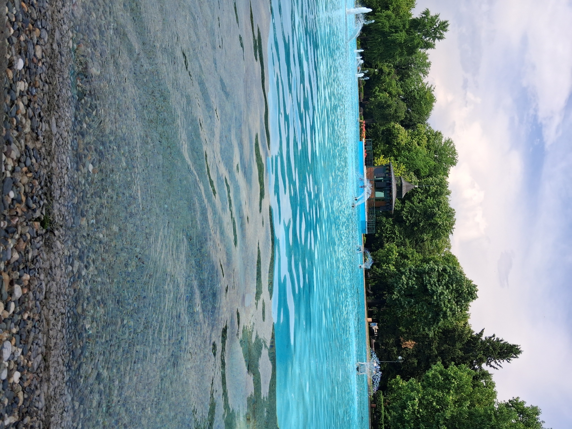 The image captures a vibrant and serene outdoor scene, likely a large decorative pool or lake within a park, presented with the pebbly shore on the left and the horizon on the right.
The primary subject is a vast body of water, characterized by its clear, light blue hue in the deeper sections and subtle ripples across its surface, reflecting the sky and surrounding greenery. To the left, the shallow edge of the water reveals a clean bed of various-sized pebbles and stones in shades of grey, brown, and white, visible through the transparent water.
Across the water, a dense line of lush green trees forms a natural backdrop under a bright, partly cloudy sky. Amidst these trees, several man-made structures are visible: a small building or kiosk with a dark roof and lighter walls, and a smaller, possibly pavilion-like structure. Numerous water fountains are active along the far edge of the water, creating white splashes and sprays. Along this edge, some spherical decorative elements, potentially lights or planters, are also noticeable. Though indistinct, several tiny figures appear in the water and near the distant structures, suggesting the presence of people enjoying the area.
The setting appears to be a well-maintained public park or garden, with the active fountains adding to its aesthetic appeal. The clear skies with scattered white clouds indicate a pleasant, sunny day, likely in the late morning or afternoon. The overall atmosphere is peaceful and inviting, suggesting a leisurely activity. No discernible text is visible within the image.