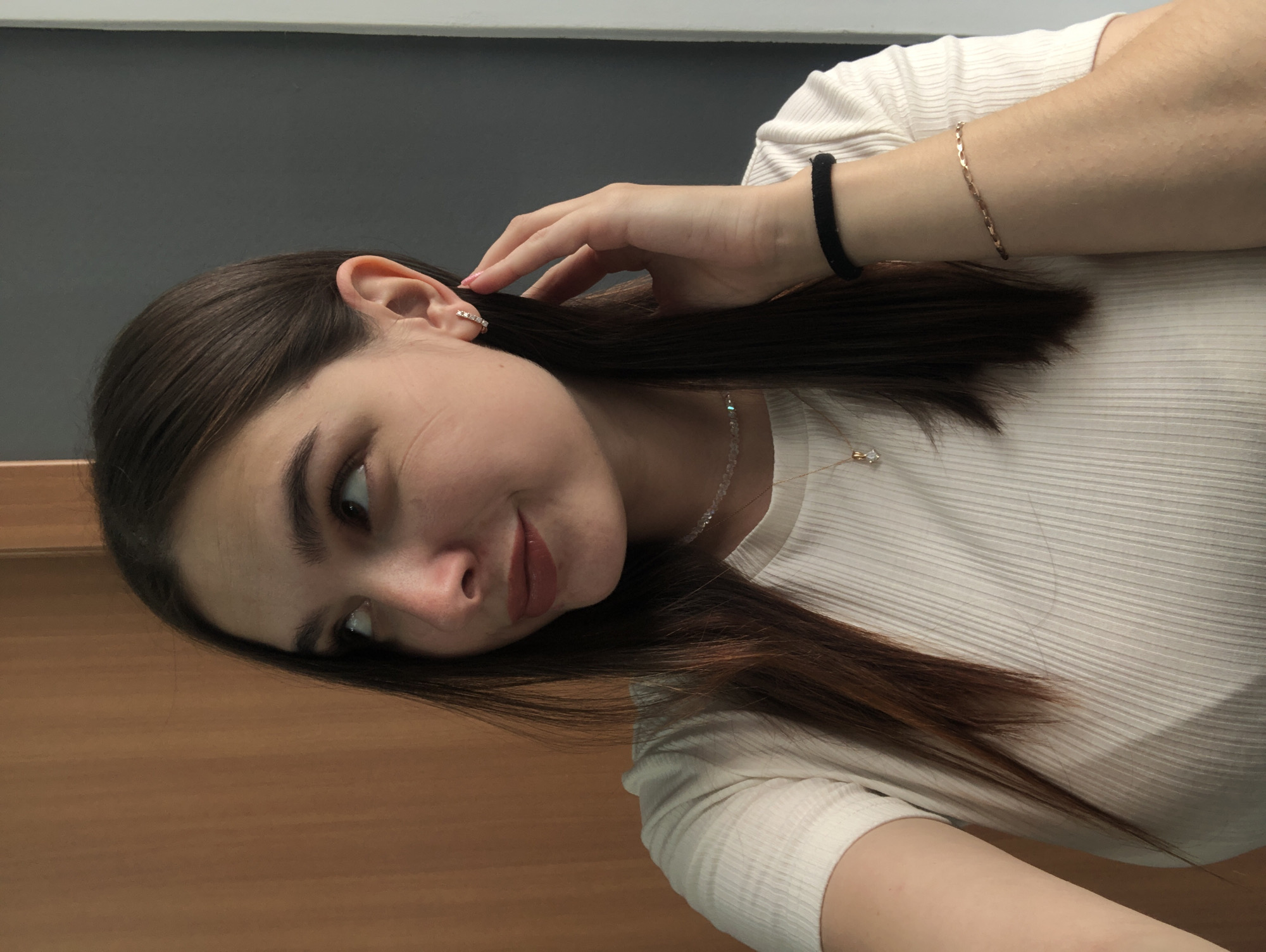 A young woman with long, dark hair is shown from the chest up, looking to her left. She is wearing a white, ribbed t-shirt, a delicate necklace with a small pendant, and small hoop earrings. A black hair tie is visible on her left wrist, and a thin gold bracelet is on her right wrist. The background consists of a light brown wooden surface on the left and a dark gray wall on the right. The lighting suggests an indoor setting, possibly a home. There are no discernible city landmarks or identifying features that would place this image specifically in Chaikovsky, Russia.