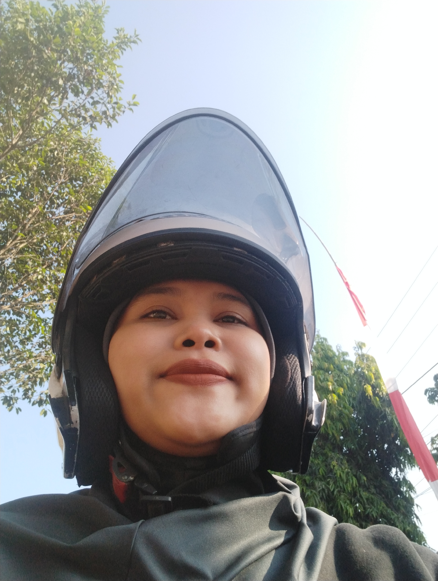 A low-angle, close-up, selfie-style shot captures a woman from the chest up, wearing a black hijab and a dark grey motorcycle helmet. The helmet's dark tinted visor obscures the top portion of her face, but her eyes, nose, and lips are visible. She is wearing dark grey clothing, likely a jacket or shirt, under the helmet. The background is a bright, slightly overexposed sky with green trees visible to the left and right. A sliver of what appears to be a red and white Indonesian flag on a pole is visible on the far right, suggesting the location is in Gambirono, Indonesia. The lighting indicates it is daytime, possibly late morning or early afternoon, with the sun high in the sky.
