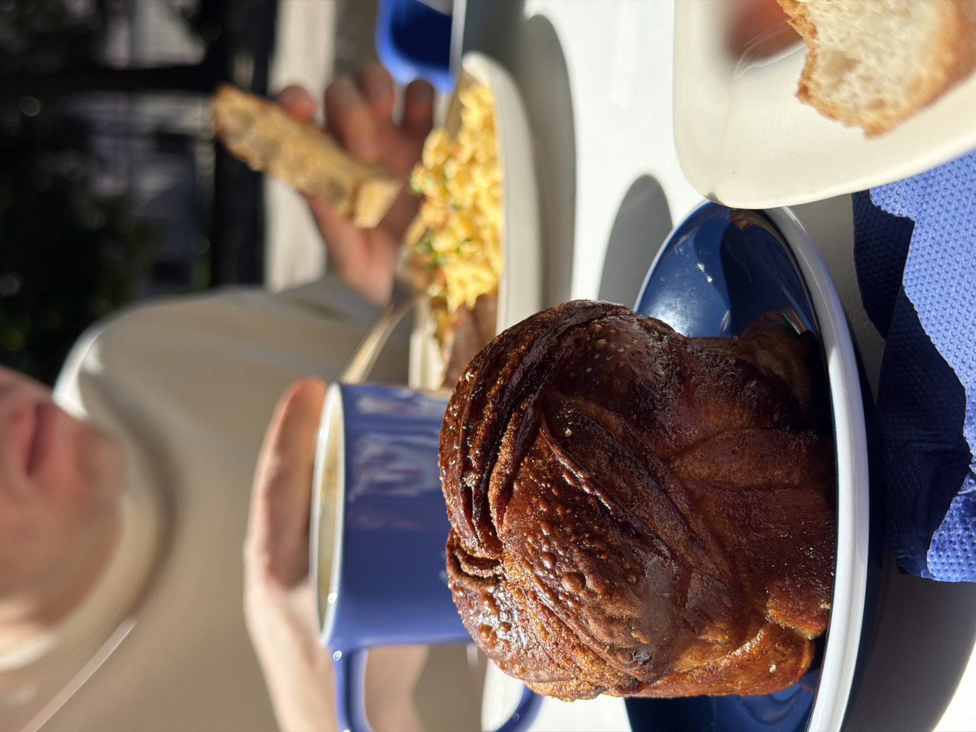A close-up, shallow depth of field shot captures a breakfast scene on a table, likely outdoors due to the sunlight and shadows. In the foreground, a rich, dark brown, intricately swirled pastry sits on a small blue plate. To its right, a light-colored square dish holds a slice of toasted bread. In the middle ground, a vibrant blue mug contains a frothy beverage, possibly coffee or latte. Behind the mug, a plate of scrambled eggs with visible green herbs is being partially eaten, with a fork in use. A hand in the background is holding a piece of toast. The person in the background, wearing a light-colored long-sleeved shirt, is partially visible from the chest up, with their mouth and chin in focus. The overall impression is a pleasant, sunlit meal. The presence of a blue mug, a common item in St. Petersburg cafes, and the style of pastries, suggest a cafe in the city.