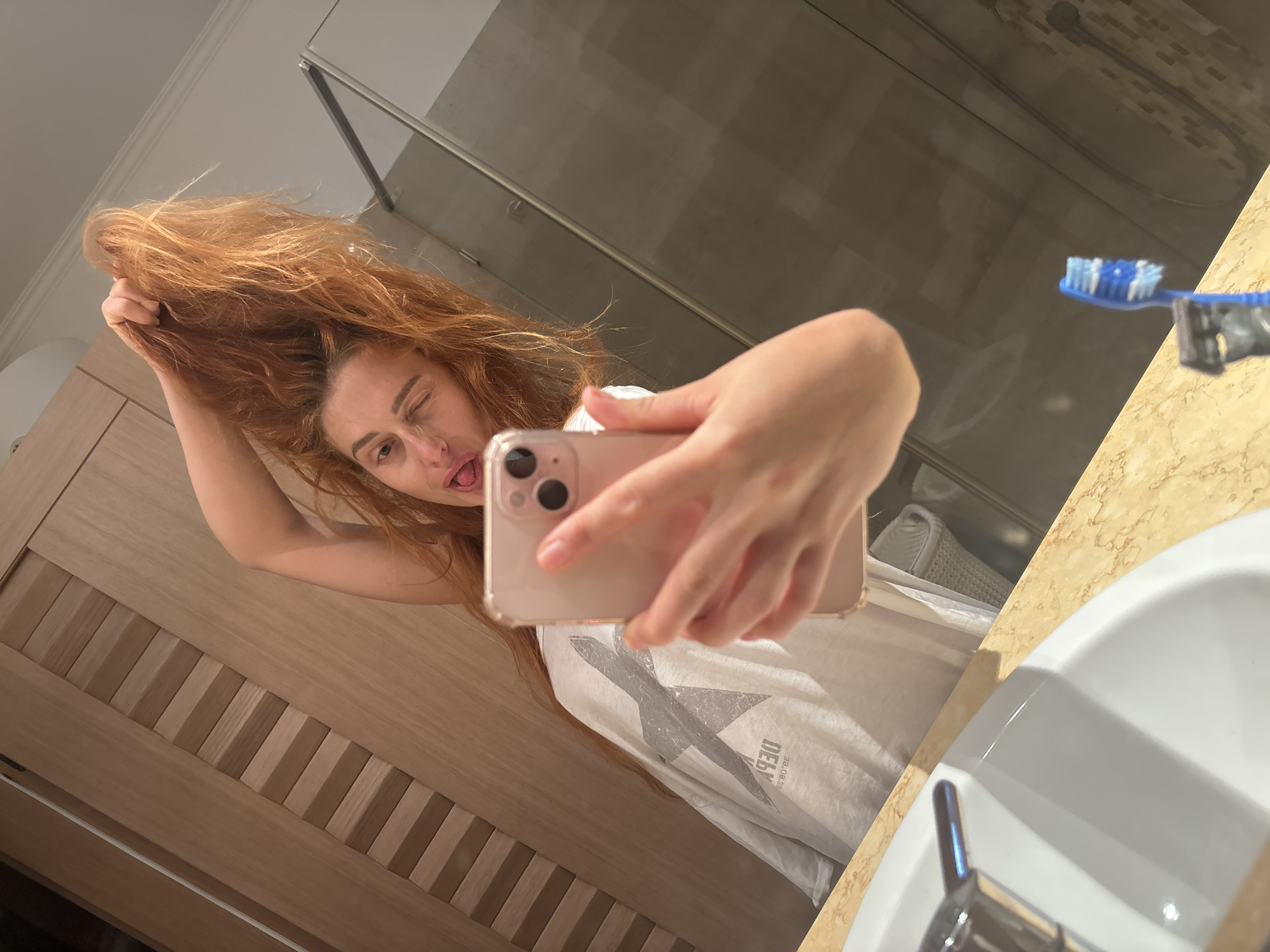 In a bathroom, a woman with long, red hair is taking a selfie in a mirror. She is holding her hair up with her left hand and sticking out her tongue while winking with her left eye. She is wearing a white t-shirt with a grey graphic print. The bathroom has wooden cabinets, a marble countertop, a white sink with a chrome faucet, a toothbrush, and a shower with a glass door. The scene is lit by artificial light. The image does not provide specific details that would allow for the identification of the city as Budva, Montenegro.