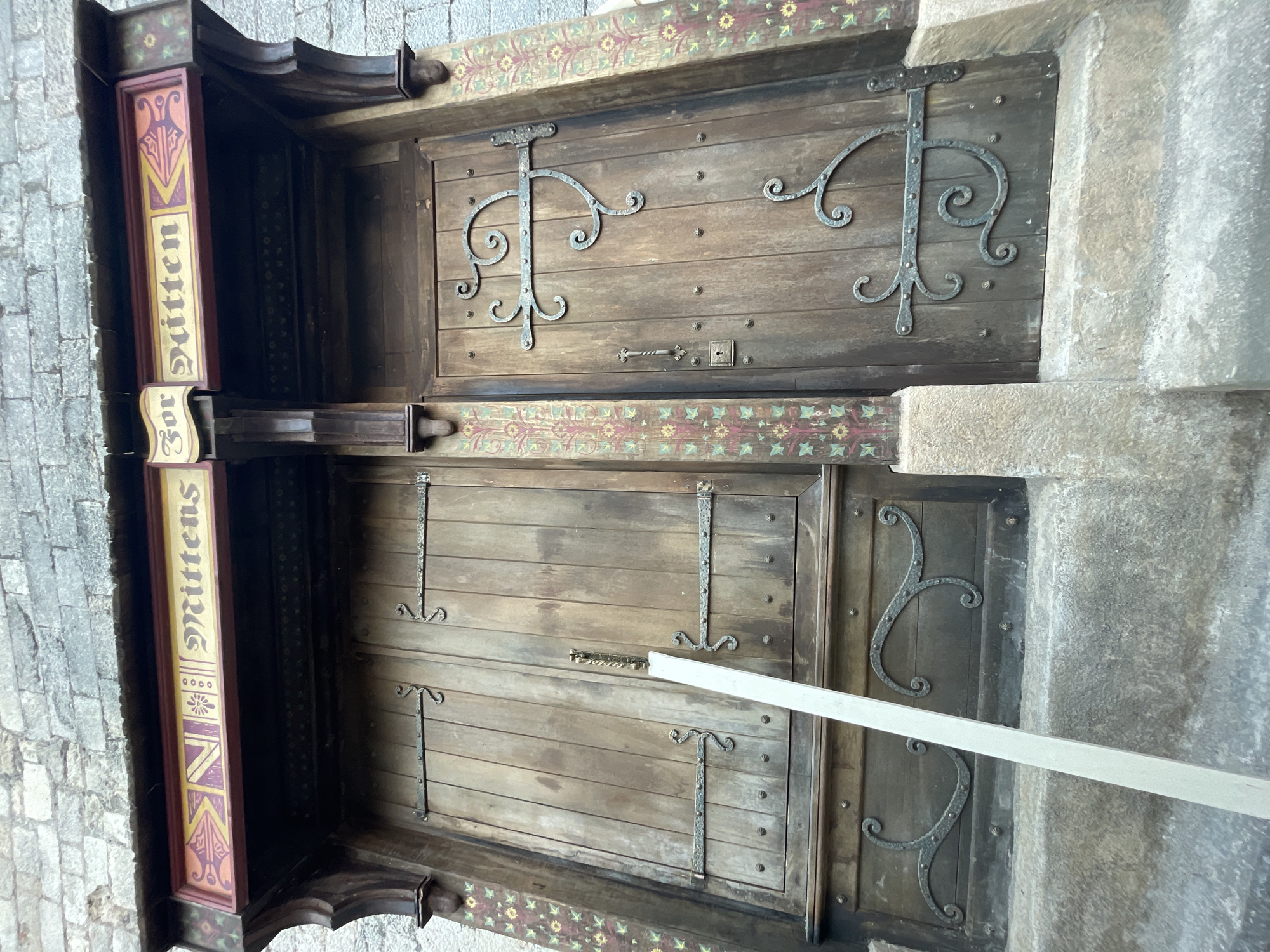 The image shows a close-up, low-angle view of a wooden double door with ornate metal hardware, set within a stone building. A sign above the door reads "Mittens" in a decorative font. The doors are weathered and appear old, with visible wood grain and metal rivets. Decorative painting in pink, yellow, and green adorns the pillars flanking the doors, and the stone facade above. A light-colored plank of wood leans against the left door, obscuring a portion of the lower half. The setting is a public space, possibly an entrance to a shop or establishment. Based on the architectural style and the name "Mittens," this likely refers to the "Mittens Shop" at the Hobbiton Movie Set in Matamata, New Zealand, not Girona, Spain.