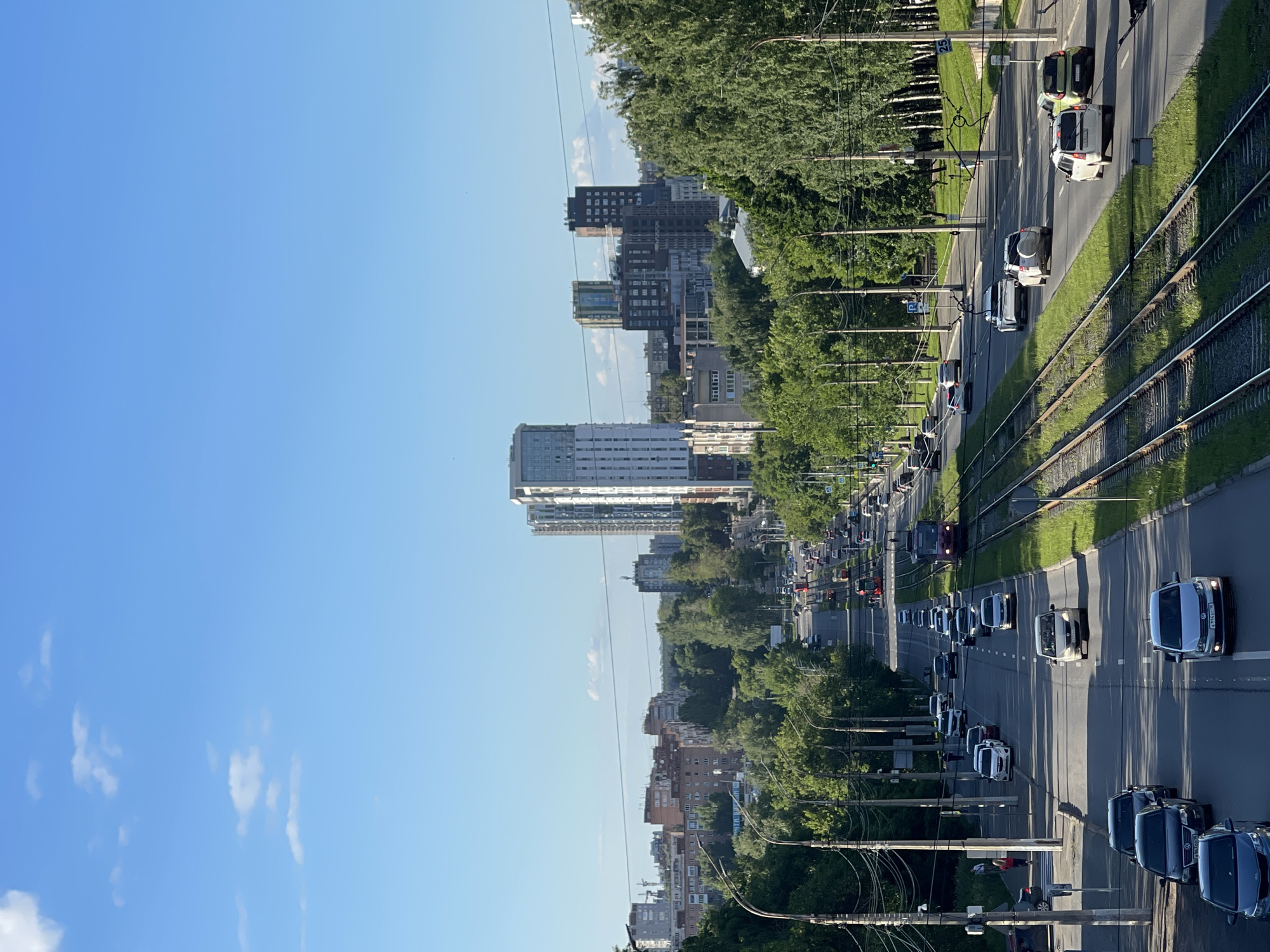 This is a medium-angle, vertical shot of a busy city street with light rail tracks. The sky is clear blue with a few wispy white clouds. In the foreground, there are multiple lanes of traffic with cars and trucks moving in both directions. The light rail tracks run parallel to the road on the right side. Several trams are visible on the tracks, one in the mid-ground and another further in the distance.
Along the sides of the road are sidewalks and numerous trees providing shade. In the background, there is a cityscape with a mix of residential and commercial buildings, including several tall modern high-rises. Power lines and streetlights are also visible throughout the scene. A few pedestrians can be seen on the sidewalks. The overall impression is a vibrant urban environment on a sunny day.