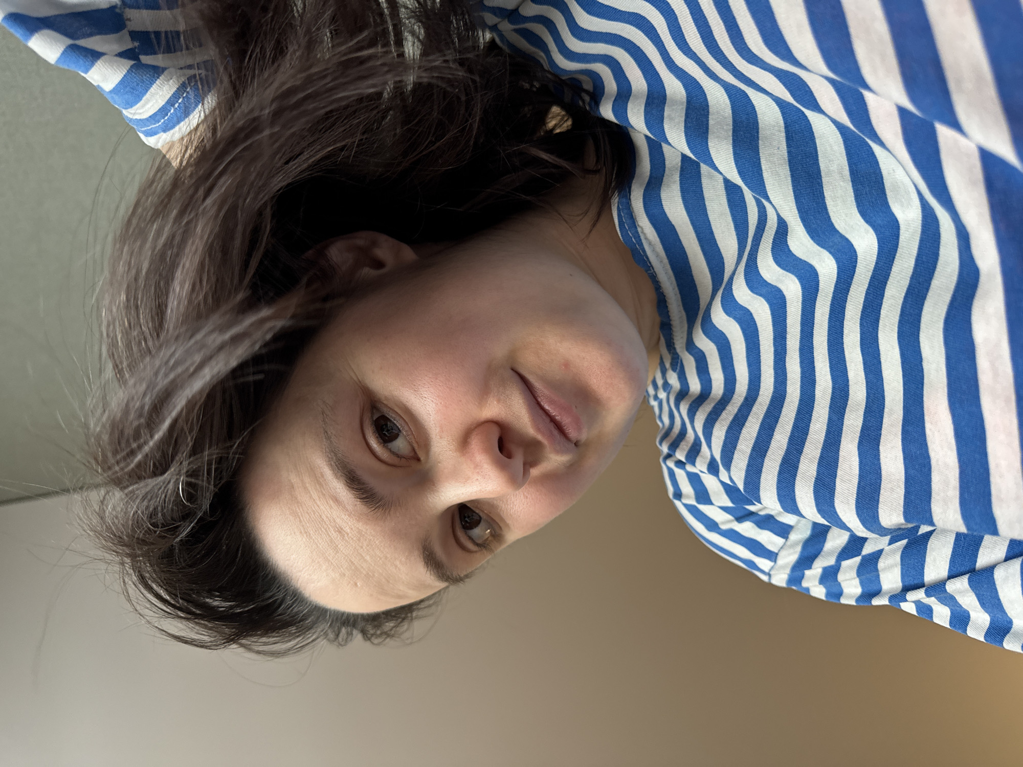 A medium shot of a woman with brown hair and light skin wearing a blue and white striped shirt. The woman is positioned in the upper center of the frame, slightly angled with her head tilted towards the left. Her eyes are open and she appears to be looking at the camera. The background is a neutral, light brown color. There are no other objects or people visible in the frame. There is no evidence to suggest the image was taken in Izhevsk, Russia.