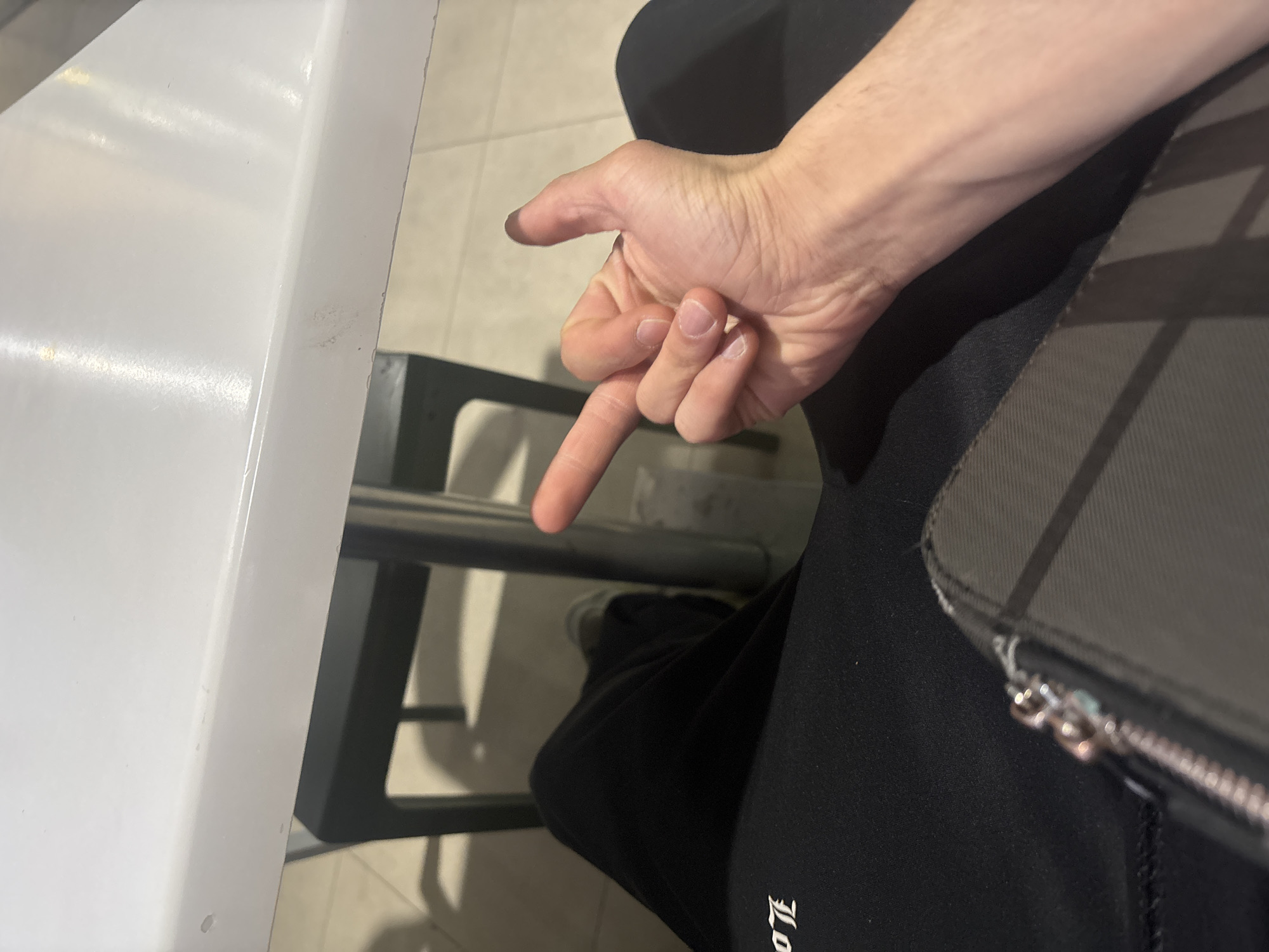 An indoor, angled close-up shot primarily features a person's right arm and hand, gesturing with an extended index finger pointing downwards and slightly forward. The thumb is also extended, while the other fingers are curled, suggesting a pointing or indicating action.
The individual is seated, wearing dark, possibly black, sweatpants or trousers. On the right side of the frame, a dark grey bag or pouch with a silver zipper is partially visible, resting next to the person's leg. Faint white stylized text, appearing to be "LO" in a Gothic-style font, is visible on the dark clothing near the bottom right.
To the left, a portion of a white, possibly laminated or plastic, table surface is visible. Beneath the table, a metal bar, likely part of the table's support or a chair, spans horizontally. The floor beneath consists of light-colored tiles. The lighting suggests an indoor setting, possibly a cafe, casual dining area, or public waiting space in Yerevan, Armenia. No specific time of day or weather conditions can be discerned. The person's face and emotions are not visible.