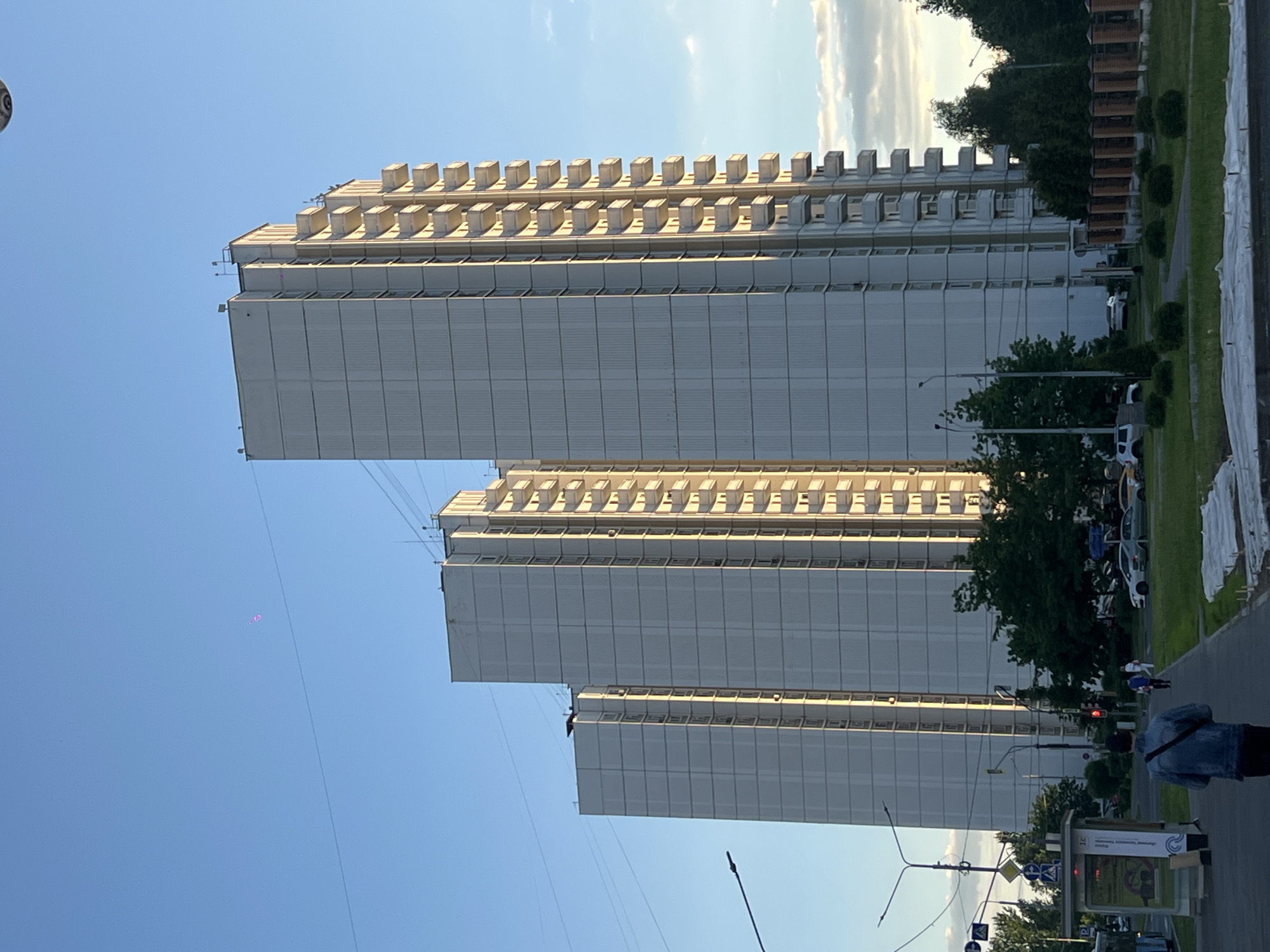 The image captures a scene in Moscow, Russia, featuring a set of three tall, white, connected buildings. These buildings, arranged vertically from the perspective of the photo, exhibit a tiered structure at the top with rectangular protrusions. The sides of the buildings are marked with vertical lines that appear to be seams or sections. In the lower-right corner, a street scene is visible, including greenery, light poles, road signs, and pedestrians. The sky is light blue with a few wisps of clouds. In the upper-left corner of the sky, a faint spot of light and an attached line are noticeable.