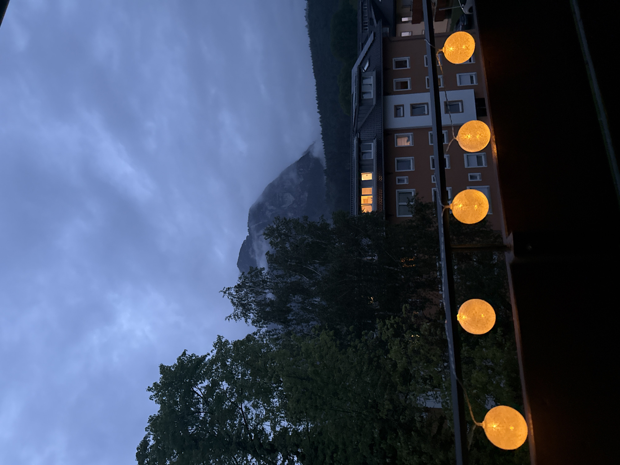 The image depicts a twilight scene from a balcony overlooking a mountainous region near Garmisch-Partenkirchen, Germany. The sky is overcast with dark, brooding clouds, obscuring the upper portions of a prominent, mist-shrouded mountain peak in the distance. Below the mountain, a dense, dark green forest covers the lower slopes. In the middle ground, several multi-story buildings with orange and white facades are visible. The windows of these buildings are lit, casting a warm glow against the darkening sky. In the foreground, a string of round, orange fairy lights is draped across a balcony railing, providing illumination to the immediate area. The lights are in focus, while the background is slightly blurred, creating a sense of depth. No people are visible in the frame. The overall atmosphere is one of quiet evening serenity, with a hint of the dramatic natural landscape dominating the background.