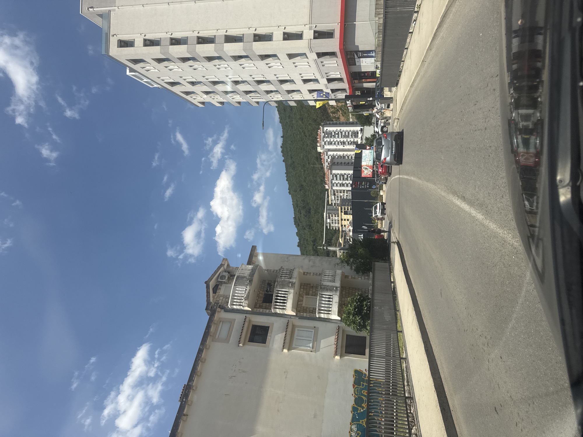 The image shows a street scene in Budva, Montenegro, on a clear day with scattered white clouds. On the left is a light-colored, multi-story building with several balconies featuring white railings. Graffiti is visible on a fence in front of this building. To the right, a modern, tall, white apartment building with numerous windows dominates the view. A red stripe runs along the bottom of this building. Cars are driving on the asphalt road, which has a white dividing line. In the background, there are other buildings and a lush green, tree-covered hill. One person is visible walking on the sidewalk near the right building. The reflection of buildings and cars is visible on the hood of a vehicle in the foreground, suggesting the photo was taken from inside a car.
