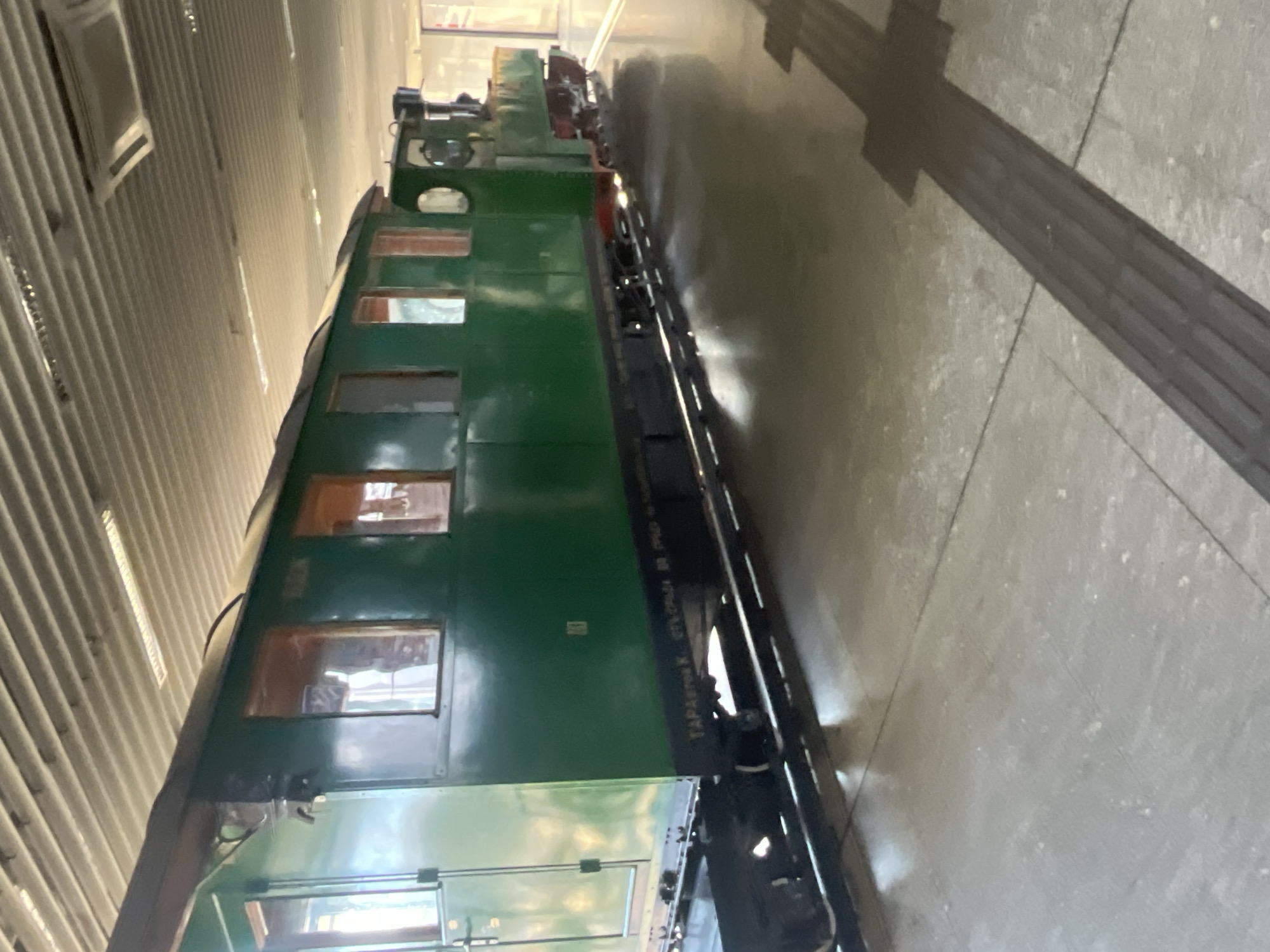 A green train car is parked indoors. The train is located in Sofia, Bulgaria. The carriage is long and has several windows. There are rails below the train car. The floor is gray. The ceiling is ribbed and angled.
