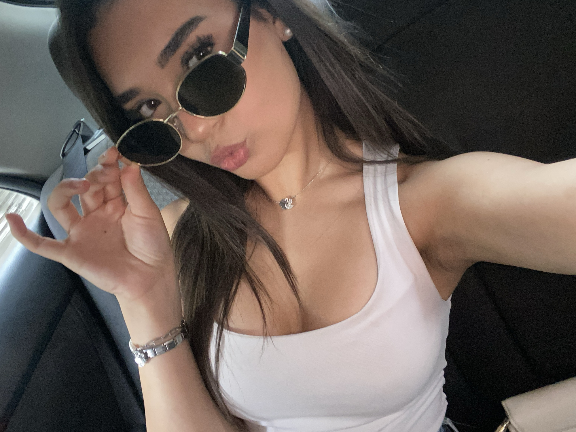 A young woman with long, dark hair is taking a selfie inside a vehicle. She is wearing a white tank top and gold-rimmed sunglasses. A silver bracelet is visible on her left wrist, and a delicate silver necklace with a floral pendant rests on her collarbone. Her right hand is raised, adjusting the sunglasses. The background is dark and appears to be the interior of a car, with a glimpse of a window. The image is likely taken in Asenovgrad, Bulgaria, based on the provided context.