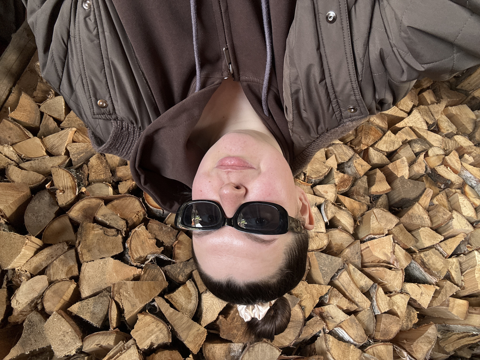 A person lies on a stack of firewood. The individual wears a brown jacket over a brown hoodie and black sunglasses. The hair is pulled back with a light-colored scrunchie. The setting appears to be outdoors. Lukintsy, Russia cannot be independently confirmed from this image.