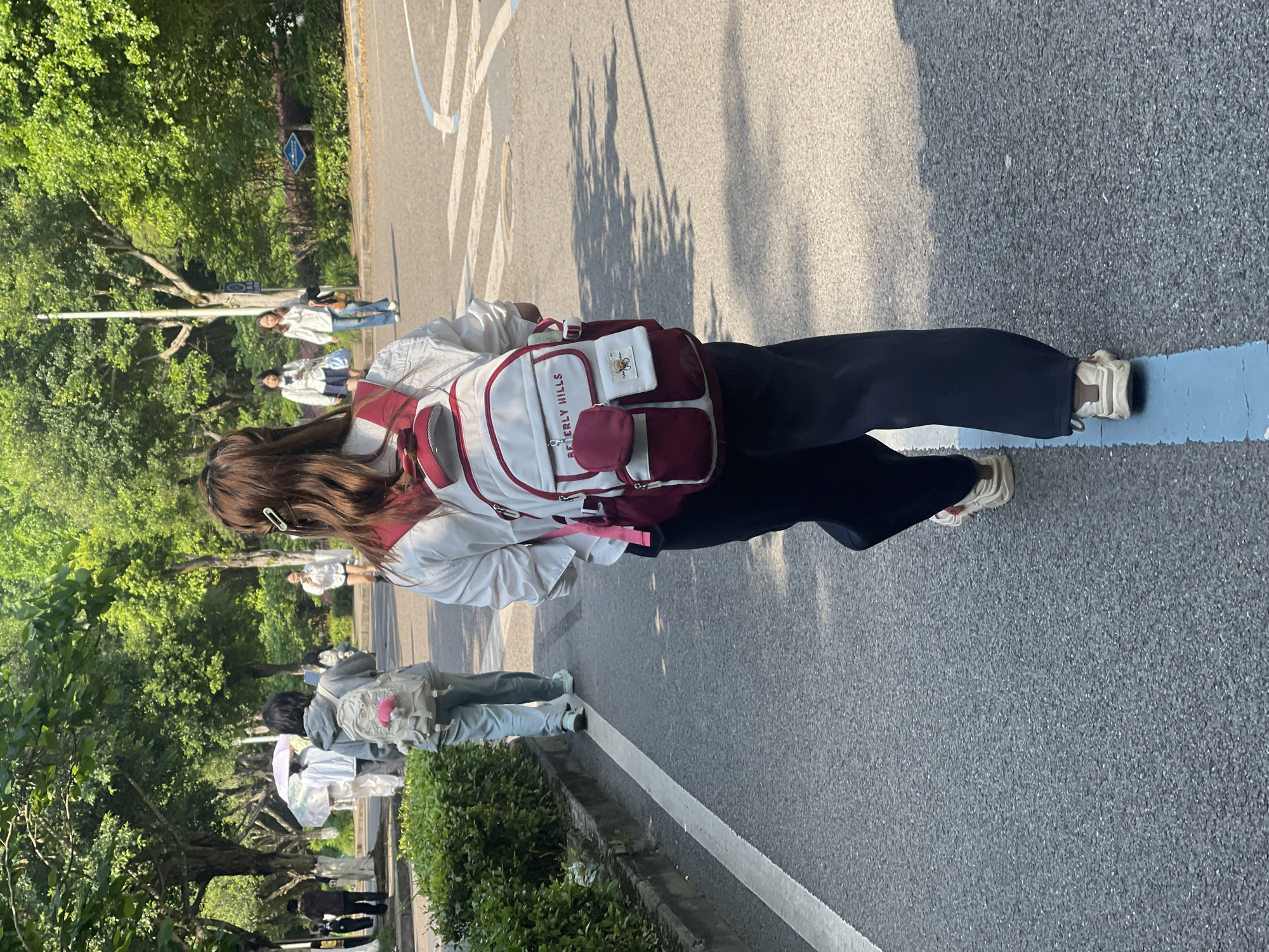 The photograph was taken in Chongqing High-tech Zone, China. The location appears to be on a road or sidewalk alongside trees and vegetation. The primary subject is a person seen from the back, wearing dark trousers, a light-colored jacket, and a backpack with a white and red color scheme. The backpack is labeled "Beverly Hills." The individual is standing on or near a blue stripe painted on the road. Other people can be seen in the background, some appearing to be students or young adults. The lighting suggests it is daytime. The environment includes paved surfaces, trees, and other greenery indicative of an urban or suburban setting with landscaping.