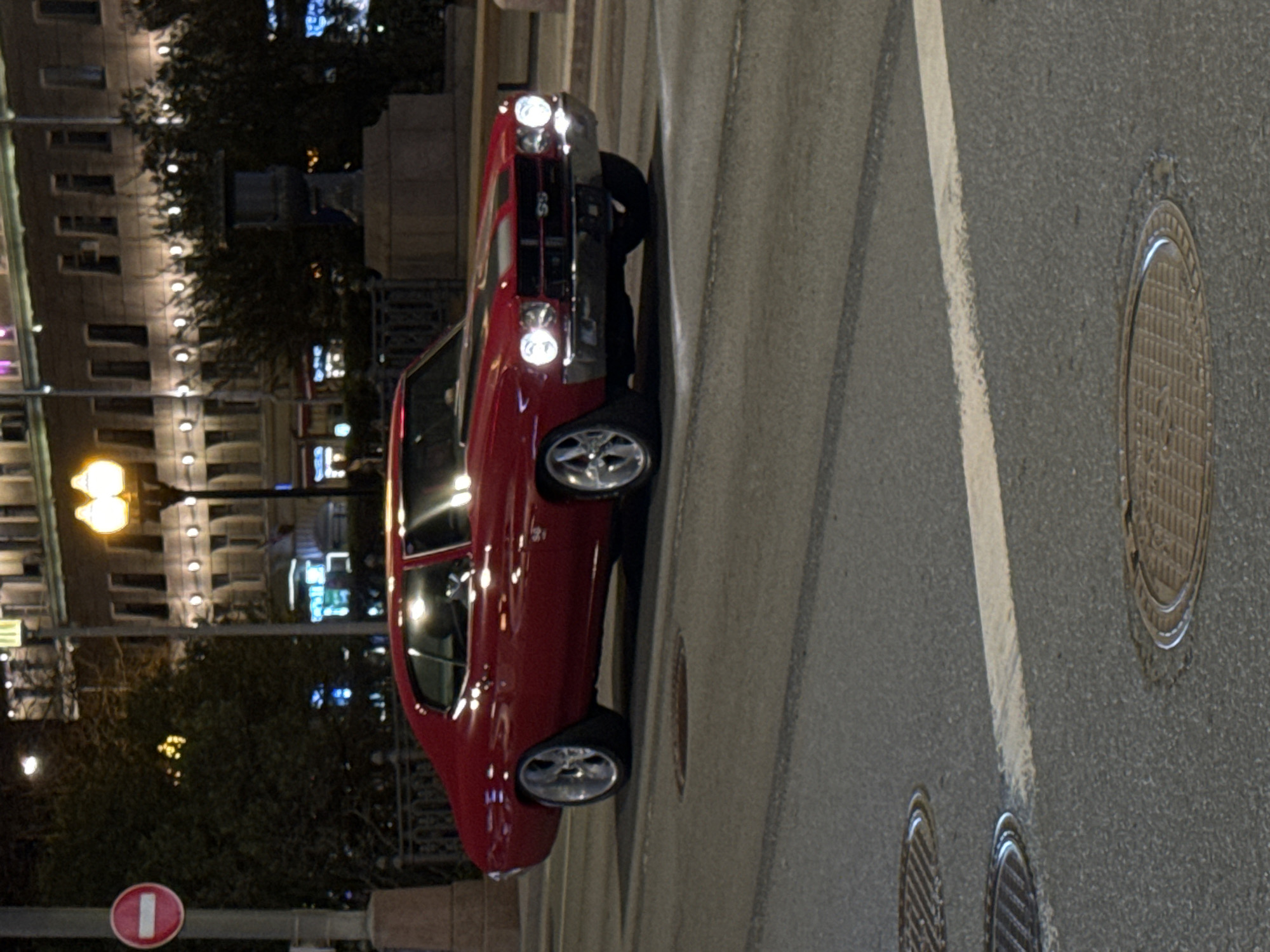 The image shows a red Chevrolet Chevelle SS parked on a city street. The car is positioned diagonally across a crosswalk, with its front end partially obstructing the pedestrian path. To the right of the car, several manhole covers are visible in the asphalt. In the background, buildings are illuminated with lights, suggesting it is nighttime. Based on the architectural style and street signs visible, the location is likely Moscow, Russia.