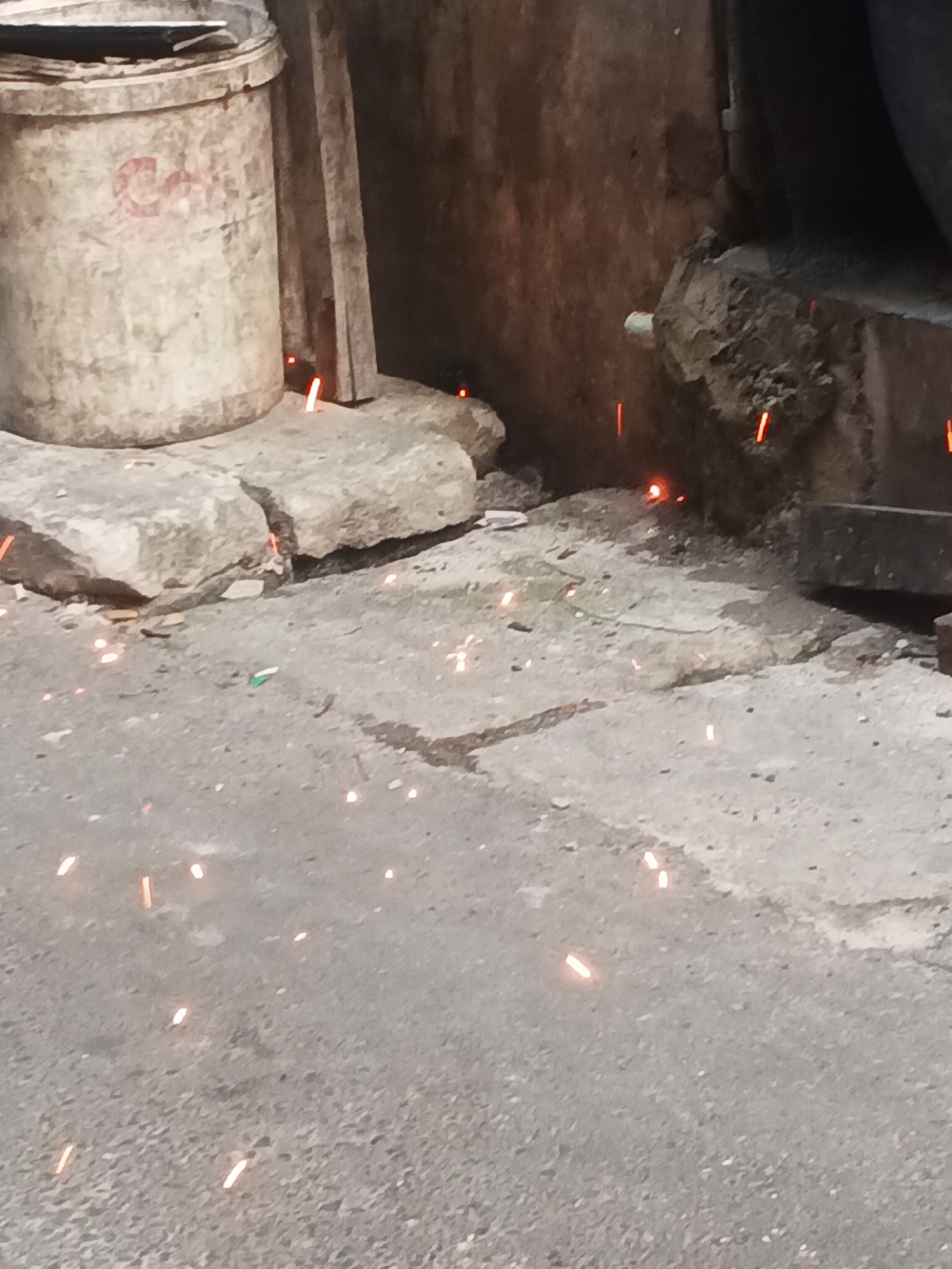 The image depicts an outdoor scene, likely a street or alleyway, in West Jakarta, Indonesia. In the foreground, a rough, grey asphalt surface is covered with numerous small, bright orange sparks, suggesting recent welding or similar hot work. A few larger pieces of concrete or stone are scattered across the ground.
To the left, a large, off-white cylindrical container, possibly a bucket or drum, is visible. It has some faded red markings on its surface. Behind the container, a vertical wooden plank leans against a textured, dark wall.
The background features a rough, uneven concrete or stone wall with visible cracks and debris. More sparks are scattered around the base of this wall and on the ground. In the upper right corner, a dark, curved object, possibly part of a larger structure or piece of machinery, is partially visible. The overall impression is of an industrial or construction area, possibly undergoing repair or demolition.