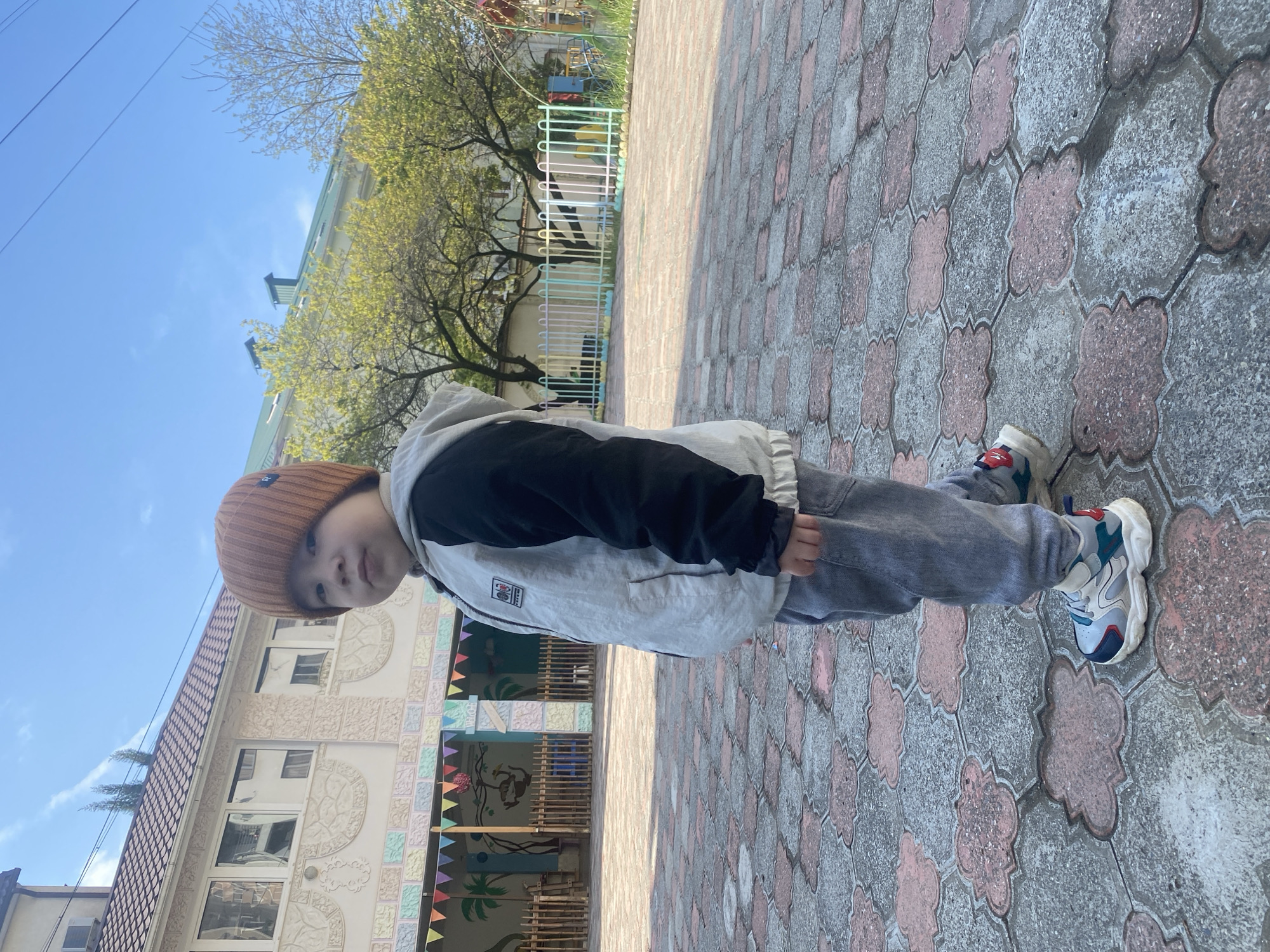 A young child is standing on a paved area with interlocking gray and pink patterned blocks. The child is wearing a brown knit hat, a gray and black jacket with a hood, gray jeans, and white, blue, and red sneakers. Behind the child is a light-colored building with windows and decorative panels, adorned with colorful flags and murals. A tree with budding green leaves is also visible. Based on visual elements such as architecture and landscaping, it is likely that the image was taken in Sochi, Russia.