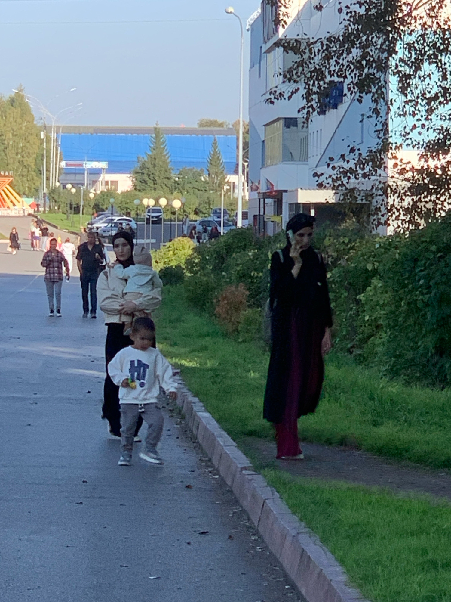 A group of people are walking on a paved path in a city park on a sunny day. In the foreground, a woman in a black hijab and long dark dress is talking on her phone while walking on the grass beside the path. Behind her, a woman in a light-colored sweater and hijab holds a baby. To the left of the women, a young boy in a white sweatshirt and gray pants walks on the paved path. In the background, several other people are walking in the park. A line of parked cars is visible further in the background, along with trees and buildings, including a large blue building. The scene appears to be in Prokopyevsk, Russia, based on the context provided.