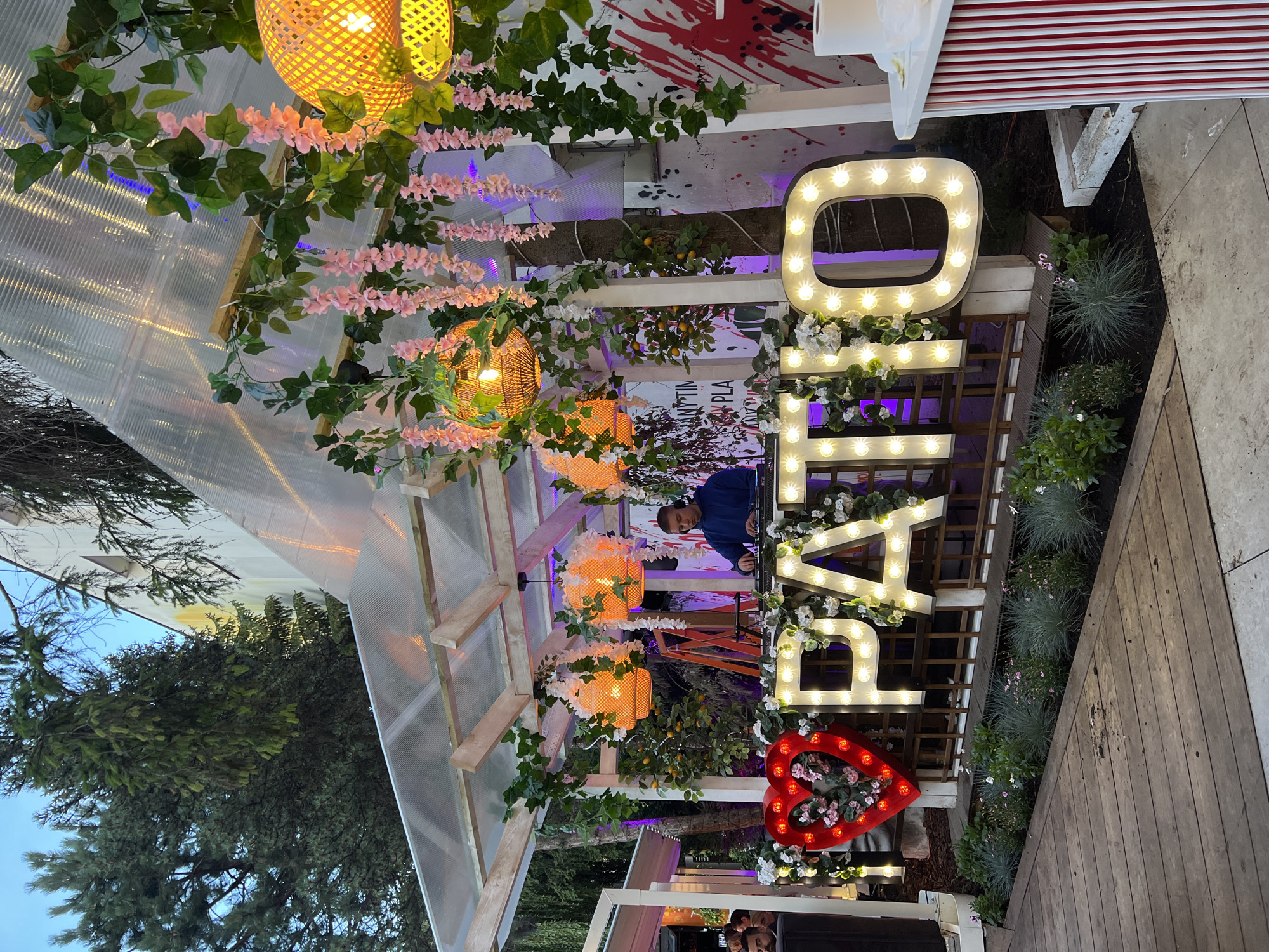 A DJ is seen at a turntable, wearing headphones and a blue hooded sweatshirt. He is positioned behind a wooden counter decorated with illuminated letters that spell out "PATIO." A large, illuminated red heart, also adorned with flowers, is to the left of the letters. The scene appears to be an outdoor patio or event space, likely at a venue named "Patio," possibly located in Białystok, Poland, given the context. The patio is covered by a translucent roof and decorated with hanging floral garlands and woven lamps. The background shows trees and a building.
