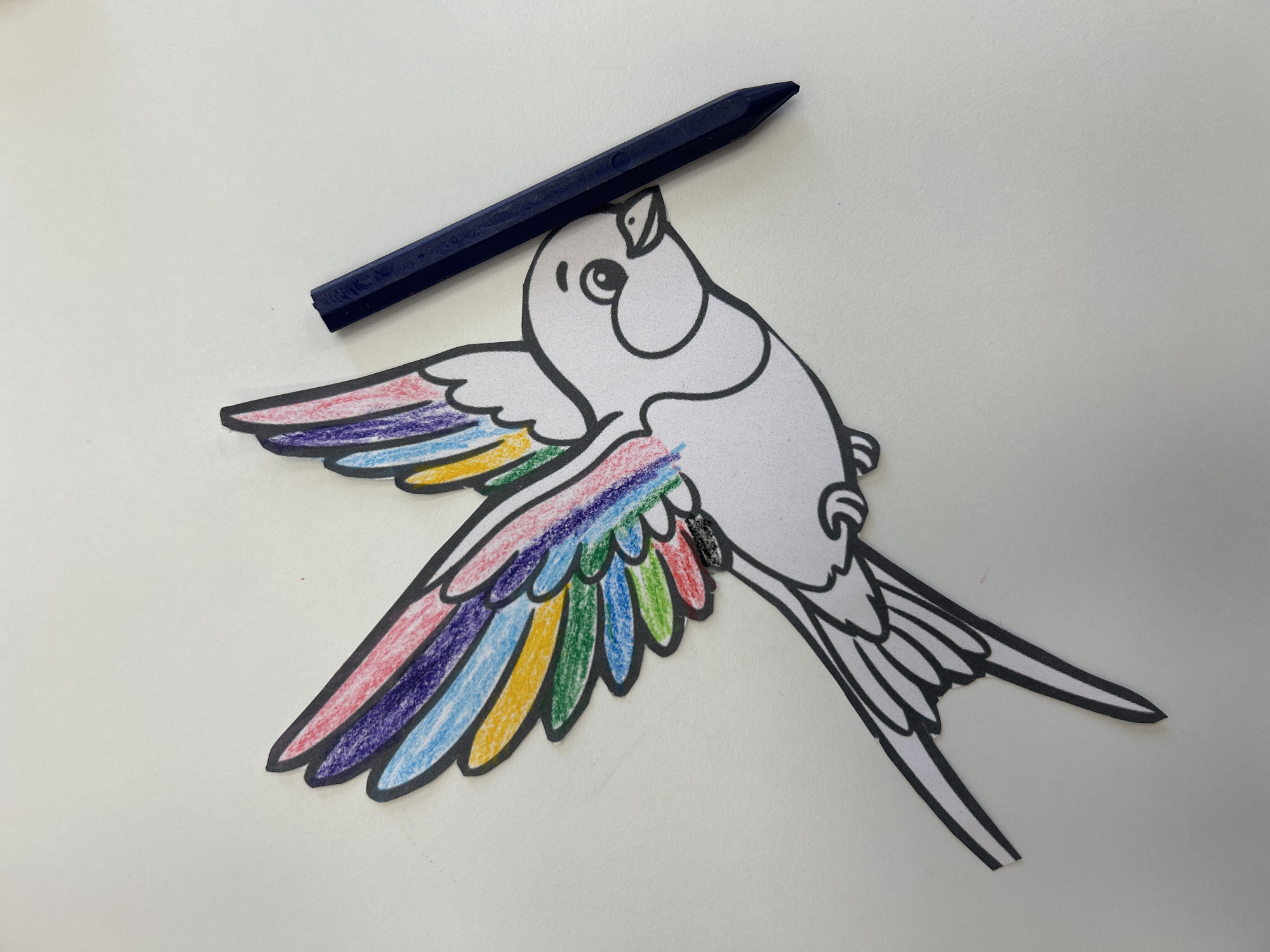 An overhead shot captures a white paper cutout of a bird with colorful wings, positioned on a white surface. The bird is depicted in mid-flight, with its wings spread and its tail fanned out. The wings are adorned with a spectrum of colors, including red, pink, purple, blue, green, and yellow. A dark blue crayon rests beside the bird, appearing to have been used to color its wings. The scene is set against a plain white background, suggesting an indoor setting, possibly a desk or table. There is no indication of the location being in Khimki, Russia.