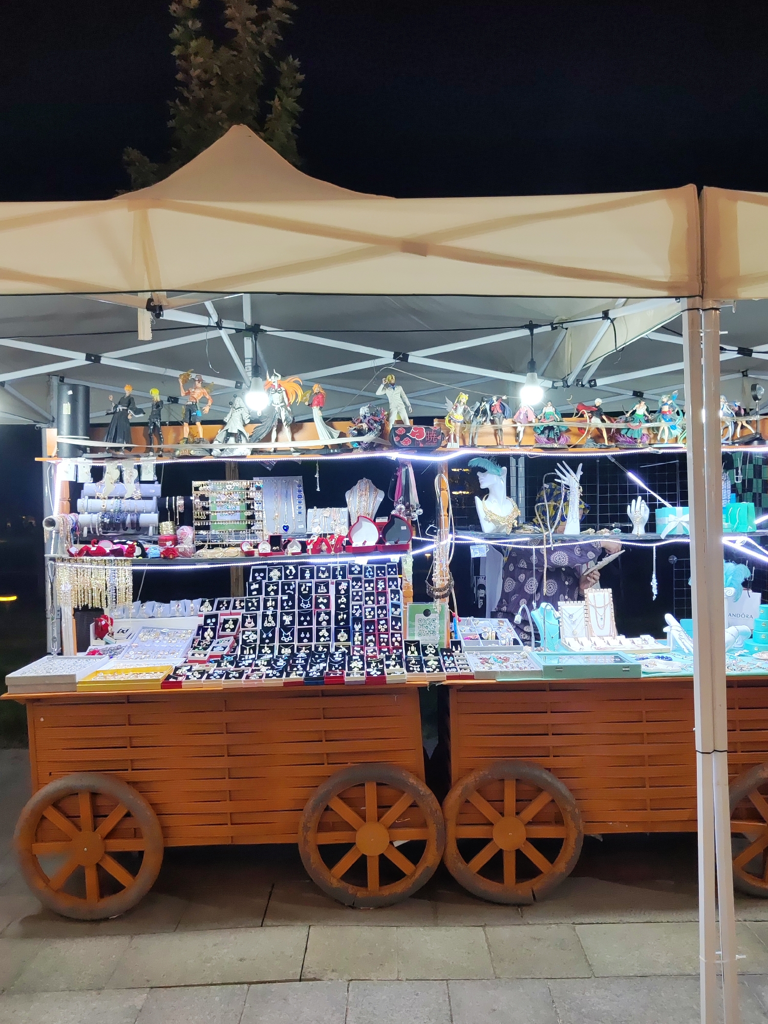 This image depicts a nighttime scene of a market stall, likely a jewelry and figurine vendor, set up under a white tent. The stall is constructed on two wooden carts with large, spoke-like wheels, giving it a whimsical, wagon-like appearance. The carts are filled with various items displayed on shelves and trays.
On the top shelf, numerous anime-style figurines are arranged. Below this, multiple shelves are laden with jewelry, including necklaces, bracelets, rings, and earrings, organized in display cases and on stands. The jewelry is illuminated by strings of white LED lights that run along the shelves, creating a festive glow. Some of the jewelry displays feature mannequins or busts to showcase necklaces and other adornments. There are also display stands shaped like hands and a mannequin head adorned with jewelry. The background is dark, suggesting it is night, with the faint silhouette of trees visible behind the tent. The stall is situated on a paved surface, possibly a sidewalk or pedestrian area. The overall impression is that of a lively night market.
Based on the style of the stall and the commonality of such vendors in popular tourist areas, this scene is highly likely to be located in the vicinity of Amir Timur Square or Broadway Street in Tashkent, Uzbekistan, known for their vibrant street vending culture.