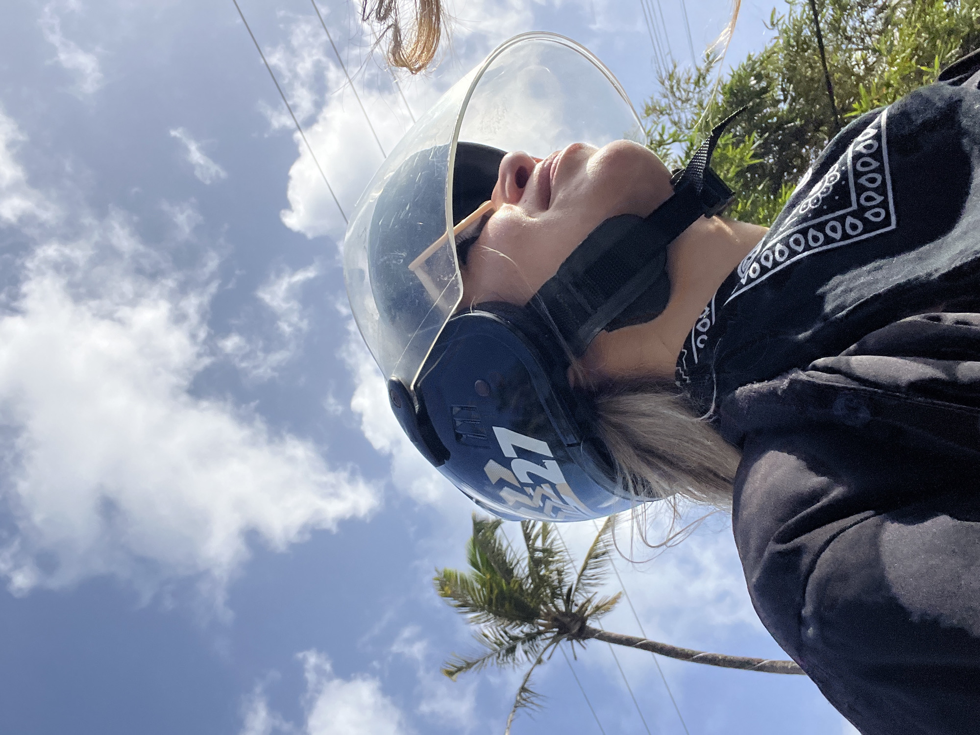 A woman is shown in a close-up, low-angle outdoor shot, wearing a black motorcycle helmet with a clear visor and a black bandana with a white paisley print. She has blonde hair and is looking upwards. The helmet has a white graphic that appears to be the number "27". In the background, a palm tree is visible, along with a bright blue sky with scattered white clouds and some power lines. The scene suggests an environment in Midigama, Sri Lanka.