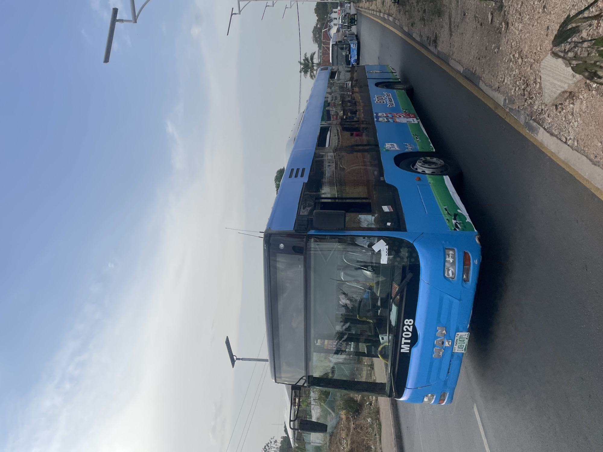 The image shows a blue MAN city bus, with the registration number "MT028," on a paved road in Jos, Nigeria. The bus is adorned with advertisements for "Enjoy Full Cream Milk" on its side. In the background, other similar buses can be seen. The surrounding environment consists of roadside vegetation and utility poles under a partly cloudy sky. The road is bordered by a painted yellow line.