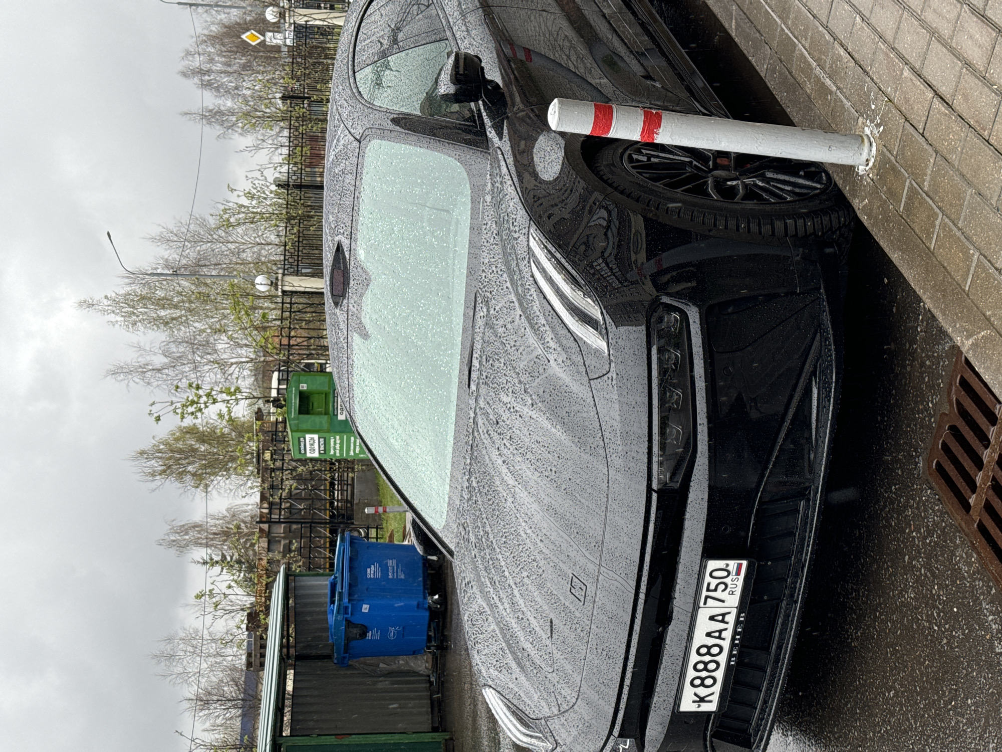 The image depicts a black car parked close to a sidewalk on a rainy day. The car's license plate reads "K888AA750 RUS BEHEB." The car appears to be next to a protective bollard, with water droplets covering the vehicle's surface.
In the background, one can see trees, a metal fence, a green recycling bin, and a blue garbage container. Based on the visible elements, it is plausible the location is Odintsovo, Russia, as indicated by the license plate containing the region code '750' that is associated with the city of Moscow.