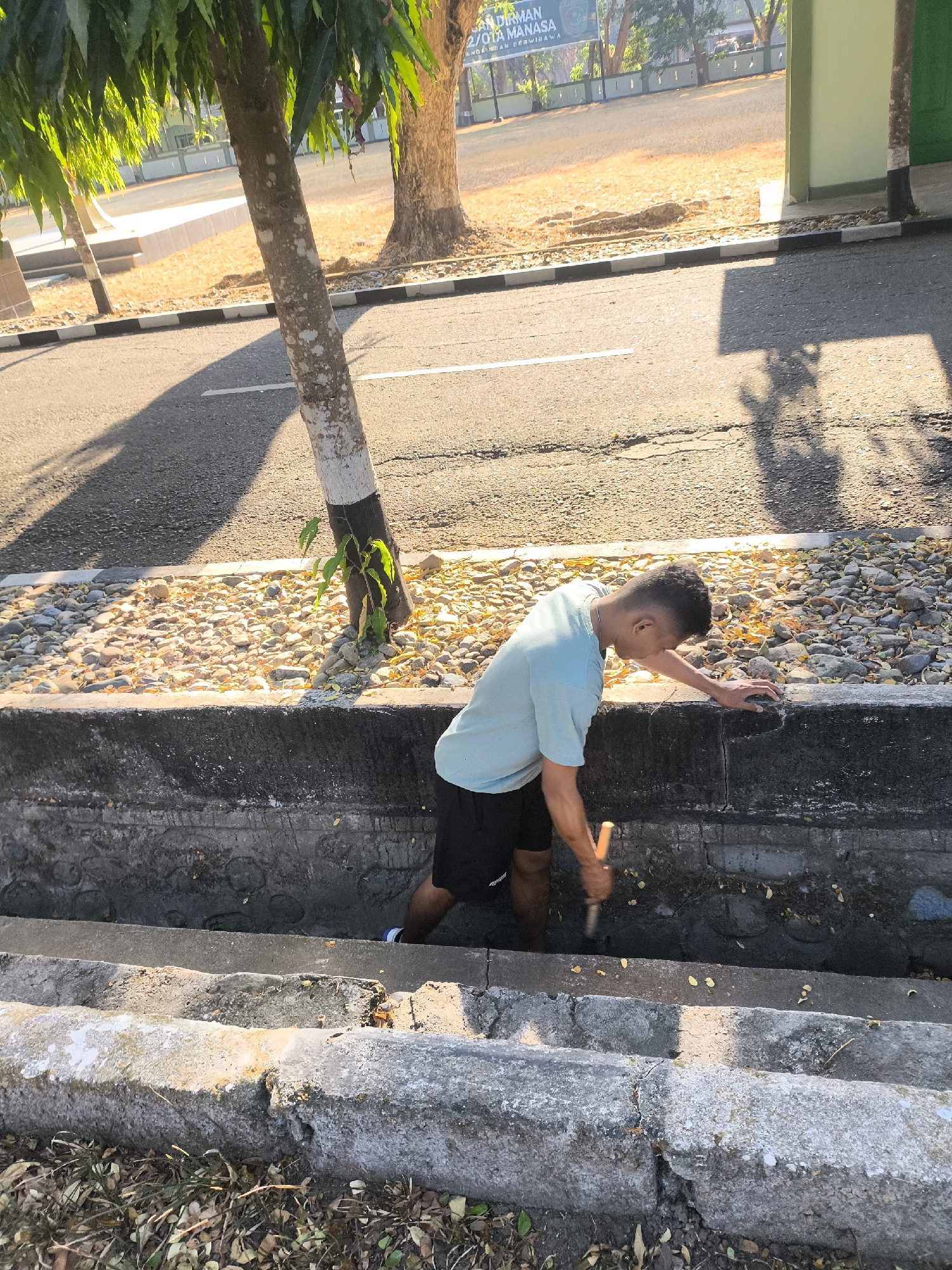 A young male, appearing to be in his late teens or early twenties, is working in a drainage ditch. He is wearing a light blue t-shirt and black shorts. The male is using a tool, possibly a shovel or trowel, to move material within the ditch. The ditch is lined with concrete, and there are stones and some dry leaves on the ground surrounding it. In the background, there is a tree with a white-painted trunk, and beyond that, a road and some buildings. The scene appears to be outdoors, under bright sunlight, casting shadows on the ground. The location is identified as Unknown, Indonesia.