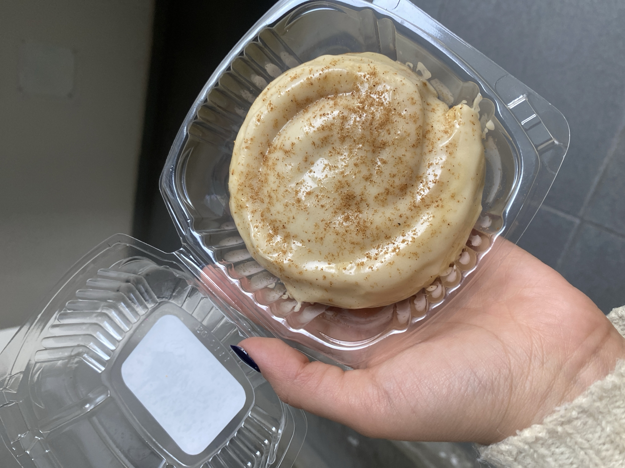 A hand holds a clear plastic container containing a frosted cinnamon roll topped with cinnamon powder. The cinnamon roll is round and the frosting is swirled. The plastic container lid is partially open and displays a white label. The background is blurry and shows a gray surface. The hand is slightly tanned and has a dark blue nail polish on the visible fingernail.