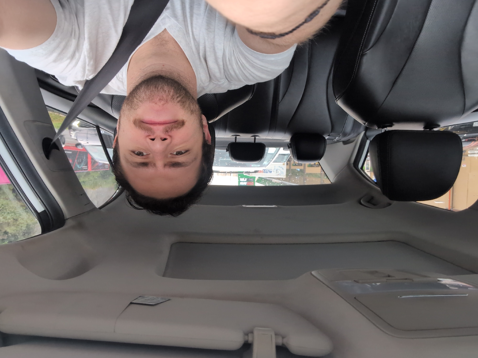 A man is inside a car in Huixquilucan de Degollado, Mexico, wearing a white T-shirt and a seatbelt. The car has black leather seats. The man is taking a selfie-style photo. Through the car windows, other vehicles and a building with the word "Autovie" are visible. The interior of the car is light grey.