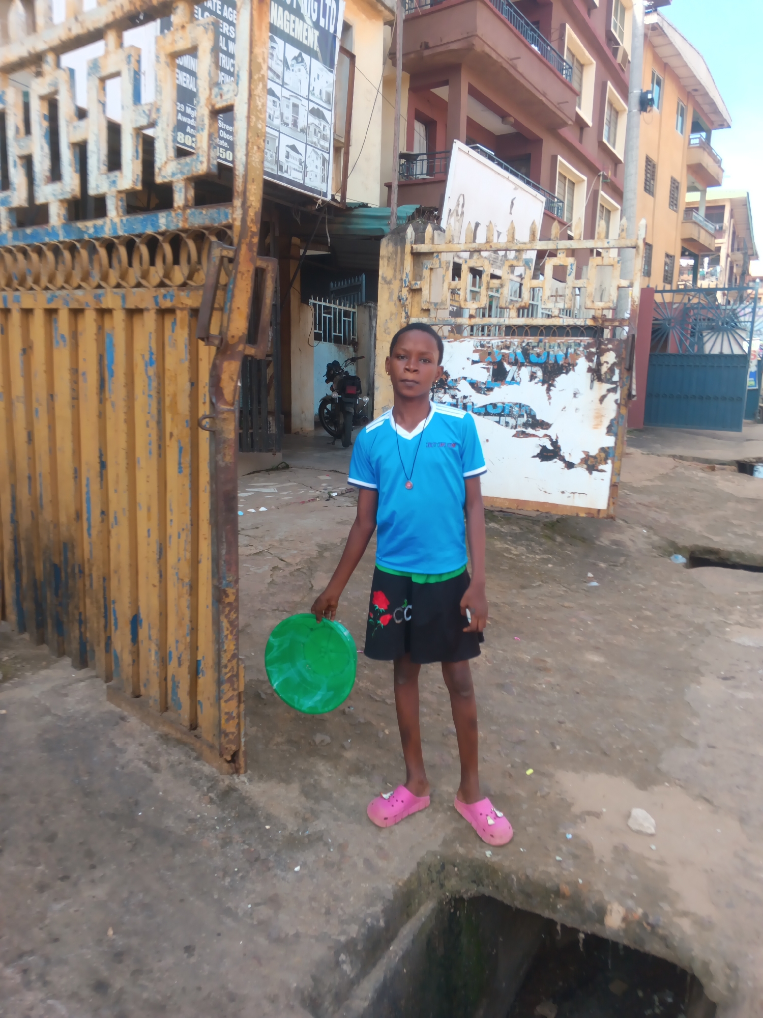 A young Black person stands outdoors in front of a rusty yellow metal gate. The person is wearing a blue t-shirt with white trim and a black skirt with red flower designs. They are holding a green plastic disc in their left hand and have pink slides on their feet. The background shows a street scene with multi-story buildings and another metal gate that is peeling paint. There is a motorcycle parked in the background, and a large, faded sign with text can be seen on the building behind the person. The ground is concrete and appears somewhat weathered, with a drainage ditch visible in the foreground. The lighting suggests it might be daytime, possibly late afternoon or early morning, as the shadows are not harsh. The overall impression is an urban setting in Nigeria. Visible text includes "MANAGEMENT" and "GENERAL".