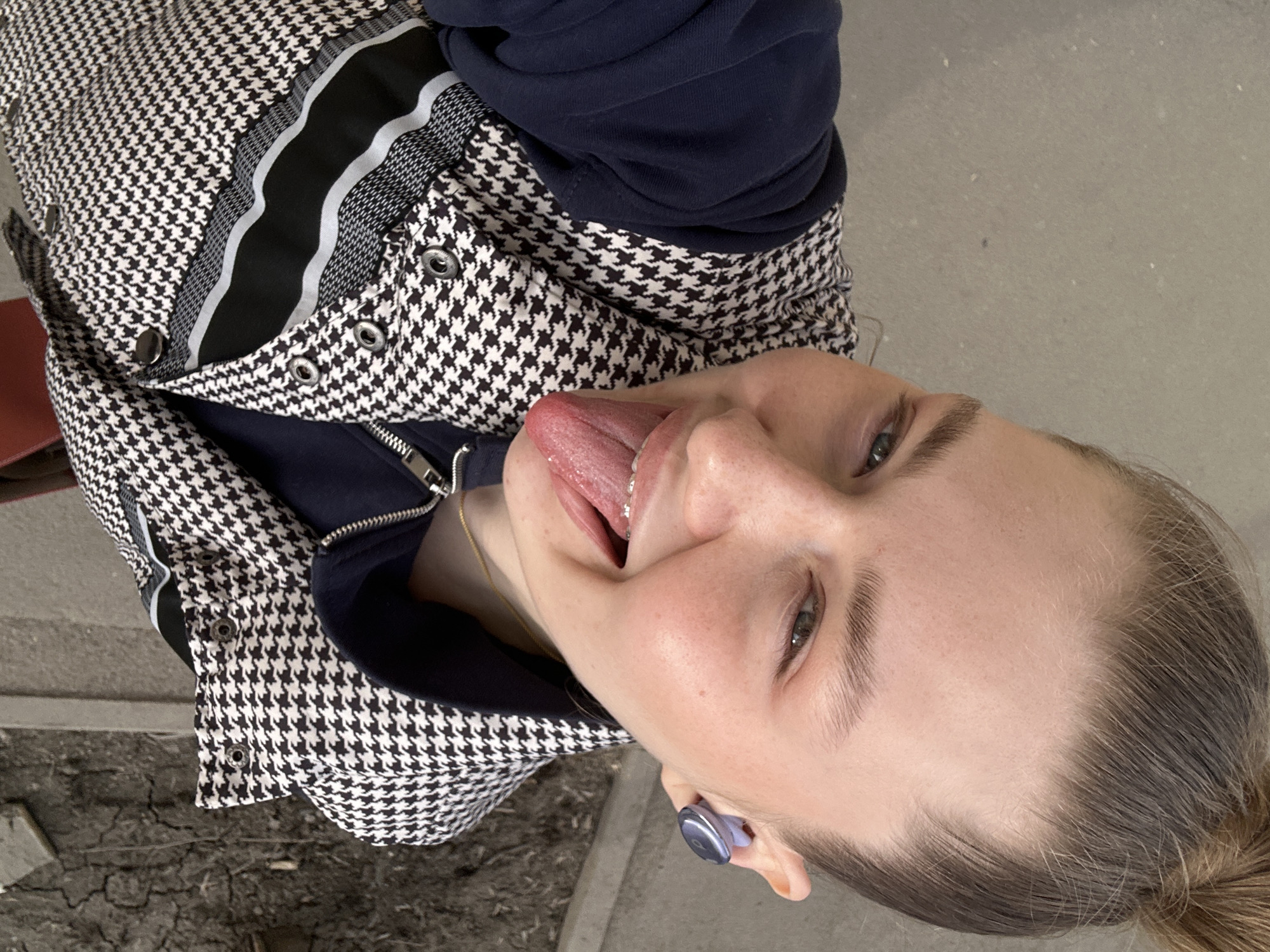 A young woman is photographed outdoors, likely in Kazan, Russia, given the image's location data. She is angled slightly to the right, sticking her tongue out, with braces visible on her lower teeth. She has fair skin, light brown hair pulled back, and is wearing an in-ear headphone in her left ear. She is dressed in a navy blue top and a houndstooth pattern jacket with a black and white stripe. The background consists of concrete surfaces, possibly a sidewalk and a wall.