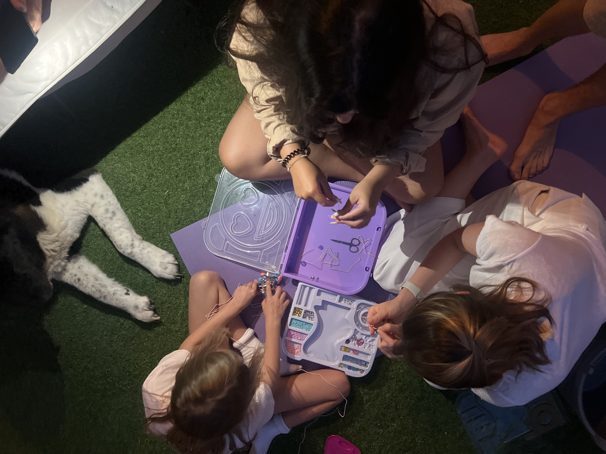 Three individuals are seated on a purple yoga mat placed on an artificial turf surface. They are engaged in making bracelets using a bead kit, which is open between them. The kit contains various colored beads, string, and a pair of scissors. One individual on the left is wearing a light pink shirt, another in the center is in a light beige shirt, and the third on the right is wearing a white shirt. A dog with black and white markings is visible in the upper portion of the image, lying down. The scene appears to be an outdoor setting, possibly a backyard or patio. The image is taken from a high-angle perspective.