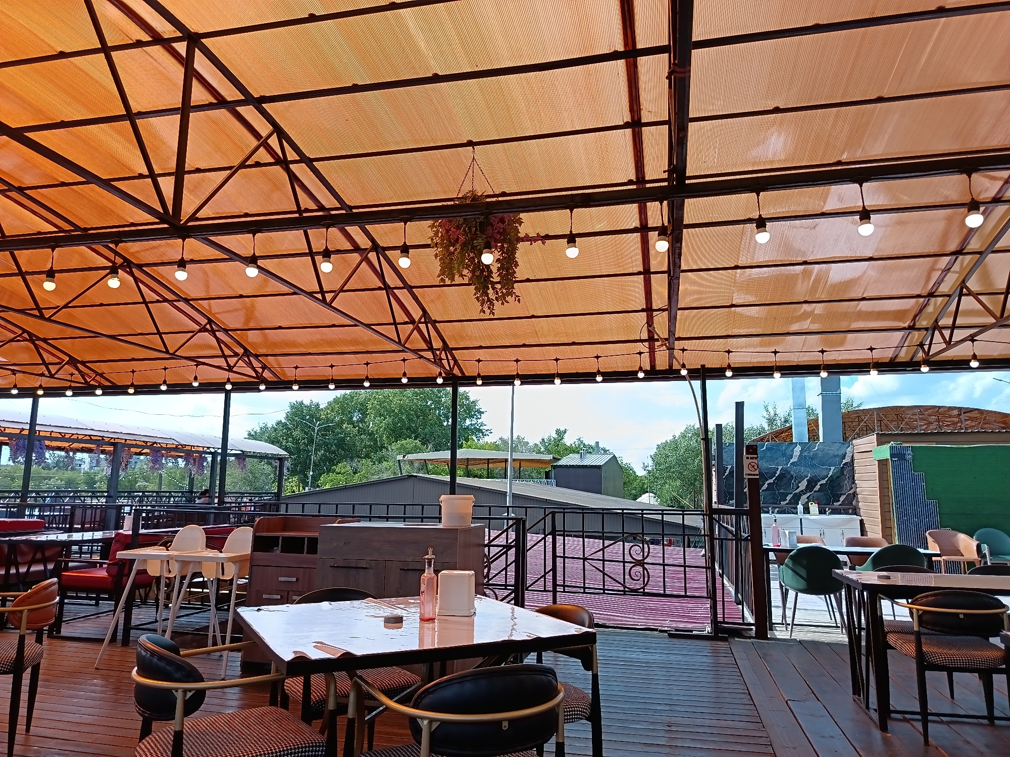 This image depicts an outdoor seating area of a restaurant or cafe, likely in Qarağandı city administration, Kazakhstan. The area is covered by an orange, translucent canopy supported by a dark metal frame. String lights are suspended from the frame, some illuminating the space. A hanging plant with green and pink foliage is visible in the center.
Several tables and chairs are arranged on a wooden deck. In the foreground, a table is covered with a white tablecloth and has a pink bottle and a white dispenser on it. Chairs with patterned upholstery surround the table. In the mid-ground, two white high chairs are placed near a wooden cabinet. Beyond these, other tables with chairs are visible, some with red upholstery.
The background shows a view of trees and buildings, suggesting an urban or semi-urban setting. A black metal railing encloses parts of the seating area. The overall impression is of a casually set outdoor dining space, possibly with a view of the surrounding environment. There are no people visible in the image.