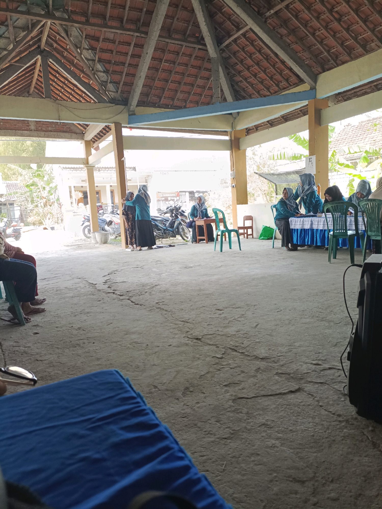 The image depicts an outdoor gathering or event taking place in what appears to be a semi-covered pavilion or open-air structure, possibly in Rancang Kencono, Indonesia. The structure has a high, exposed wooden roof with red tiles. In the background, there are buildings with tiled roofs and foliage, suggesting a residential or semi-rural setting.
Several individuals are present, most of whom are wearing headscarves, indicating a likely Muslim cultural context. In the mid-ground, a group of women are gathered around tables draped with blue cloths. Some appear to be seated, while others are standing and interacting, possibly engaged in registration, information sharing, or some form of service. Green plastic chairs are positioned around the tables.
Further to the left, two individuals are seated on chairs, facing away from the camera. One person is wearing a blue covering and the other is wearing a patterned dress. Their legs and feet are visible, with one person wearing flip-flops.
In the far left of the frame, a blue mat or tarp is visible in the foreground, partially obscuring the ground. A pair of eyeglasses is resting on the mat. To the right of the frame, a black electronic device, possibly a speaker, is situated on the ground.
The ground surface is concrete, showing some cracks and imperfections. Motorcycles are parked in the background, indicating transportation is present. The overall scene suggests a community event or activity taking place in a relatively informal, open-air setting.