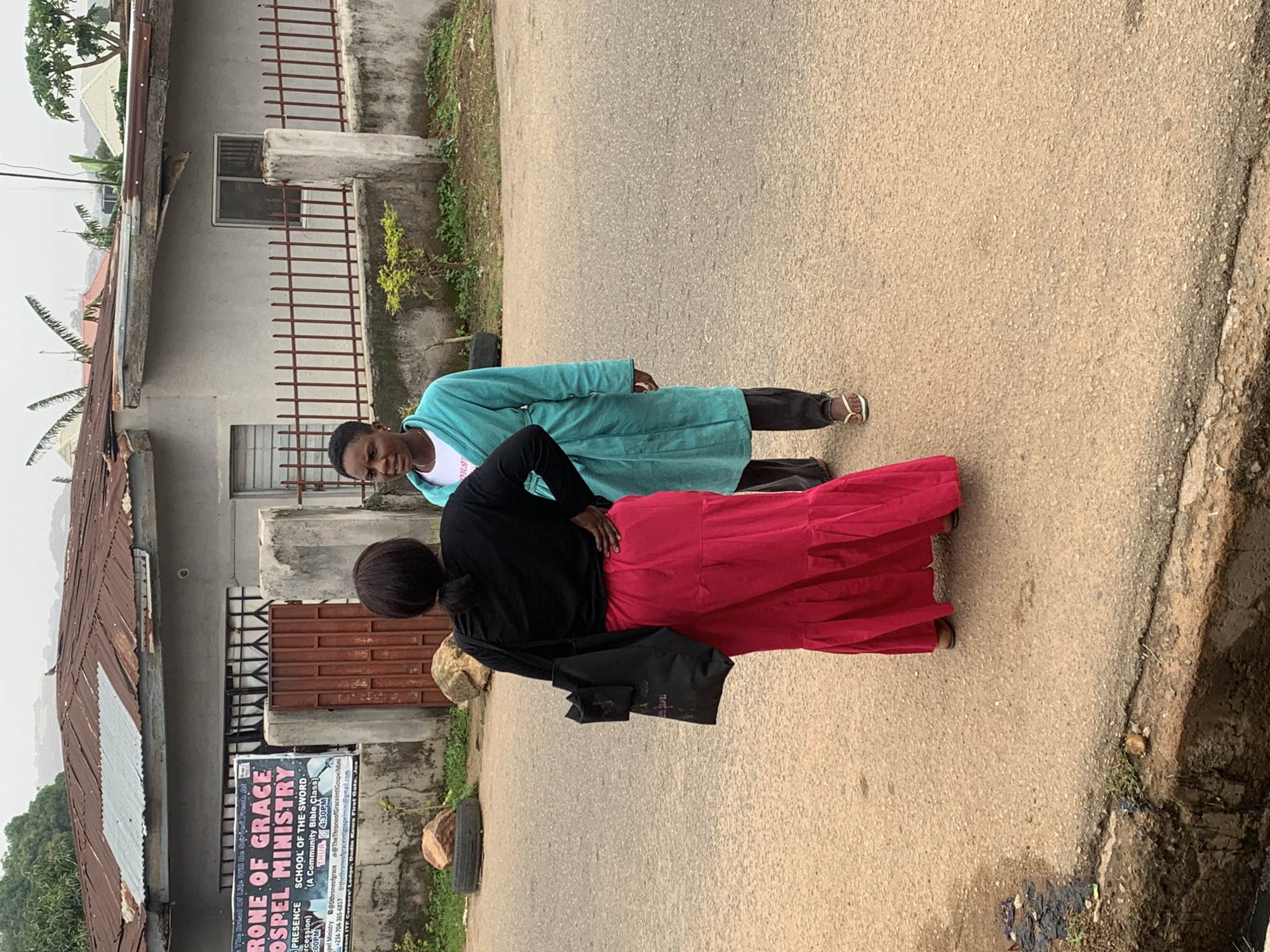 Two individuals are standing on a paved road in front of a building in Jos, Nigeria. The person on the left is wearing a black long-sleeved shirt and a long, bright pink ruffled skirt, with a black bag slung over their shoulder. They are facing away from the camera, with their left hand on their hip. The person on the right is wearing a teal-colored robe over dark pants and white flip-flops, facing the other person and looking at them. They are standing in front of a light-colored, single-story building with a rusty corrugated metal roof. A red metal gate is visible to the left of the building, and a banner with text is attached to the wall. A metal fence runs along the front of the property. The ground is an asphalt road with a concrete drainage ditch along the left side, containing some debris. The background shows more buildings and vegetation under an overcast sky.