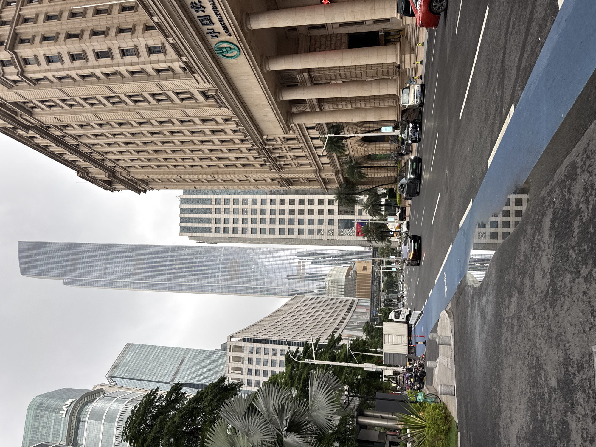 A street view in Tianhe District, China, shows a modern urban scene with towering skyscrapers and a multi-lane road. The sky is overcast.
On the right side of the image is a large, light-colored building with a classical facade featuring rows of windows and large columns. Above the entrance, there is signage with Chinese characters and "AGRICULTURAL BANK OF CHINA".
To the left of the classical building, other modern skyscrapers dominate the background. One is a very tall, slender glass skyscraper, while another has a patterned facade with vertical lines. A building with a curved, green glass facade is visible further to the left.
The street in the foreground is wet, reflecting the buildings and sky. A blue-marked lane runs along the right side of the road. Several cars are driving on the road, including a dark sedan in the foreground right and a white van further back.
On the far left, there is a sidewalk area with some greenery, including palm-like plants and trees. A white truck is parked on the side of the road, and several motorcycles are also visible. The overall impression is a busy, contemporary city environment.
