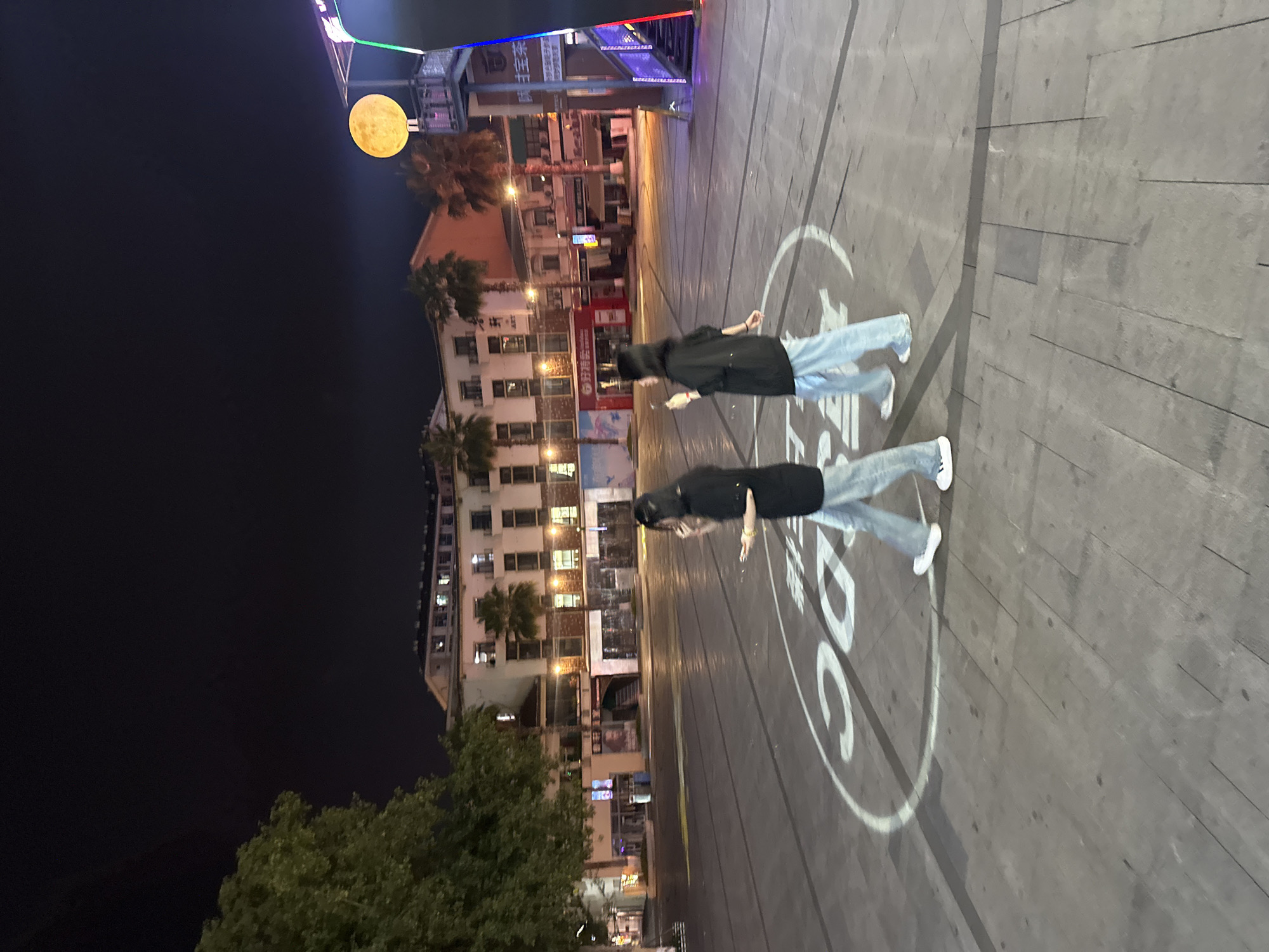 At night, two individuals walk across a paved plaza in the city of Chongqing High-tech Zone, China. The plaza features a large circular graphic with Chinese characters and the number "30" illuminated on the ground. In the background, a multi-story building with illuminated windows and palm trees can be seen. To the right, a tall structure with a large, glowing circular light resembling a moon stands beside a staircase with colored lights. The scene is dimly lit, suggesting it is late evening or early morning. The individuals are dressed casually in dark tops and light-colored pants. The plaza appears to be an open public space, possibly a pedestrian area or a town square.