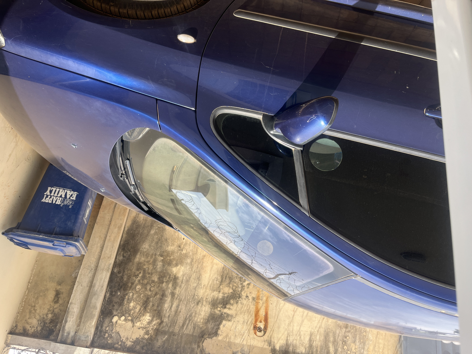 A close-up, upward-facing shot shows the front portion of a blue car, including the headlight, side mirror, and the edge of a tire. A blue trash bin with white lettering partially visible, reading "HAPPY FAMILY," sits to the left. The car is parked on a concrete surface with stains and streaks. Reflected in the car's headlight is a barbed wire fence and the sky. The city and specific location are unknown.