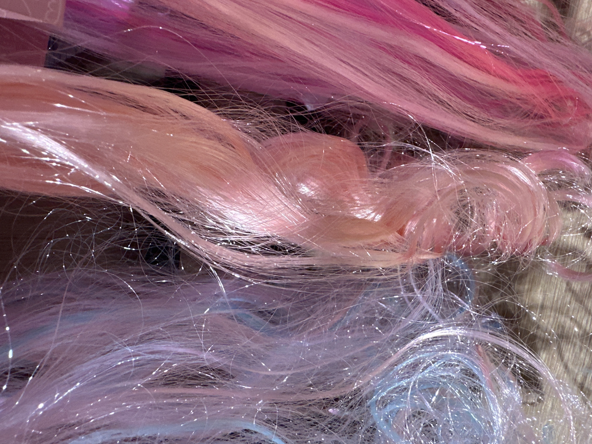 The media file is a close-up, high-angle shot of several strands of synthetic hair, likely from a wig or extensions. The hair is multi-colored, predominantly in shades of pastel pink, lavender, and light blue. Interspersed with the colored strands are fine, shimmering threads, possibly metallic or glittery fibers, which catch the light and create a sparkling effect. Some of the hair is loosely braided or twisted, creating texture and depth within the image. The background appears to be a textured surface, possibly fabric or a mat, in a neutral, earthy tone. The lighting is bright, highlighting the glossy texture of the hair and the reflective quality of the shimmering threads. There is no discernible activity or event taking place; the image focuses solely on the material itself. The overall impression is one of vibrant, playful, and possibly whimsical aesthetic, often associated with fashion accessories, cosplay, or decorative items. No text is visible in the image.