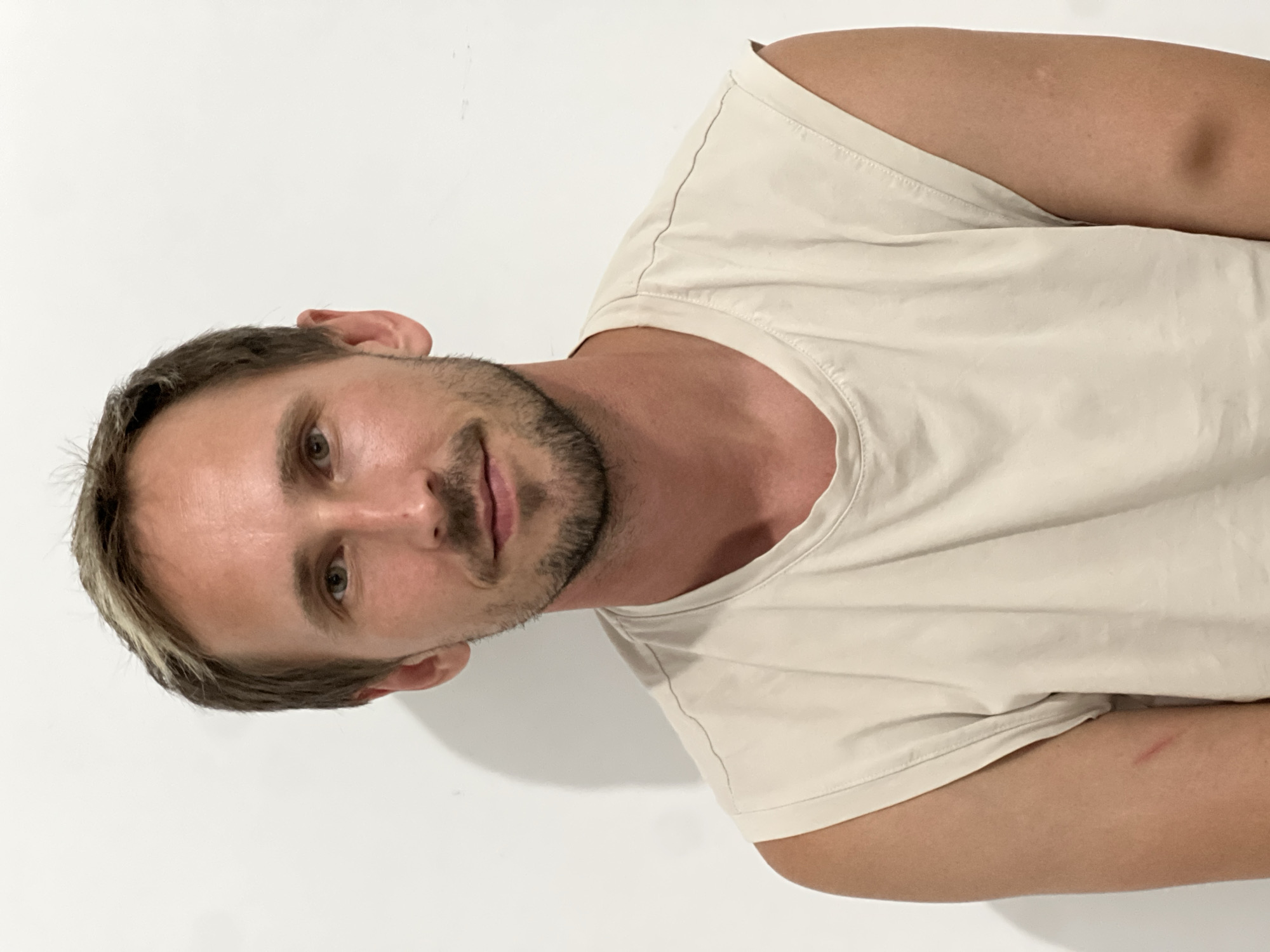 A medium shot photograph depicts a man with light skin, brown hair with some lighter streaks, and a short beard and mustache. He is wearing a sleeveless, light beige t-shirt and is seated against a plain white wall with a shadow cast to his left. The man is looking directly at the camera with a neutral expression. His left arm exhibits a faint reddish mark. The background is uniformly white and devoid of any discernible features, suggesting an indoor setting. The lighting appears to be even, with no harsh shadows except for the one behind the man. The photograph's composition is centered on the man's face and upper body. This scene does not provide any visual clues to its specific location within Kiryat Bialik, Israel, or any other city.