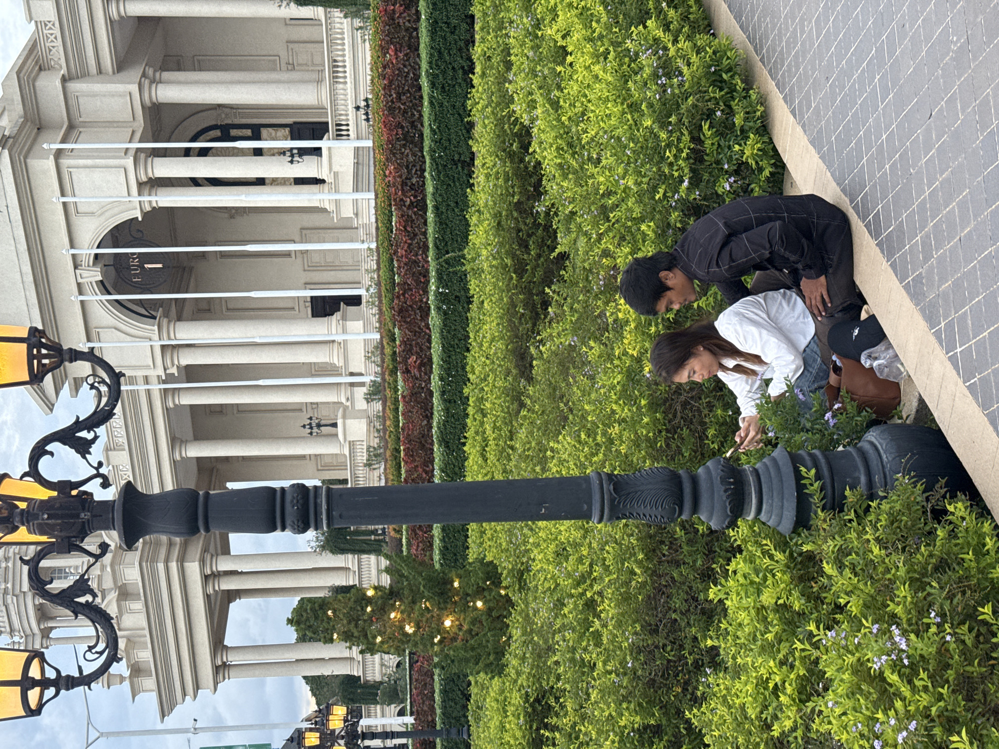 In the city of Khan Chbar Ampov, Cambodia, two individuals are seated on a raised, paved pathway adjacent to a lush green garden. The man, wearing a black checkered shirt and dark pants, sits to the right of the woman. The woman, dressed in a white shirt and blue jeans, is holding a mobile phone in her hands and appears to be interacting with it. Beside her is a brown bag. The garden features dense green foliage with scattered small purple flowers. Behind the garden, a black lamppost with ornate details stands prominently, and further back, a large, ornate building with classical architecture, characterized by columns and arches, is visible under a cloudy sky. In the background, a small, decorated evergreen tree is illuminated with fairy lights.