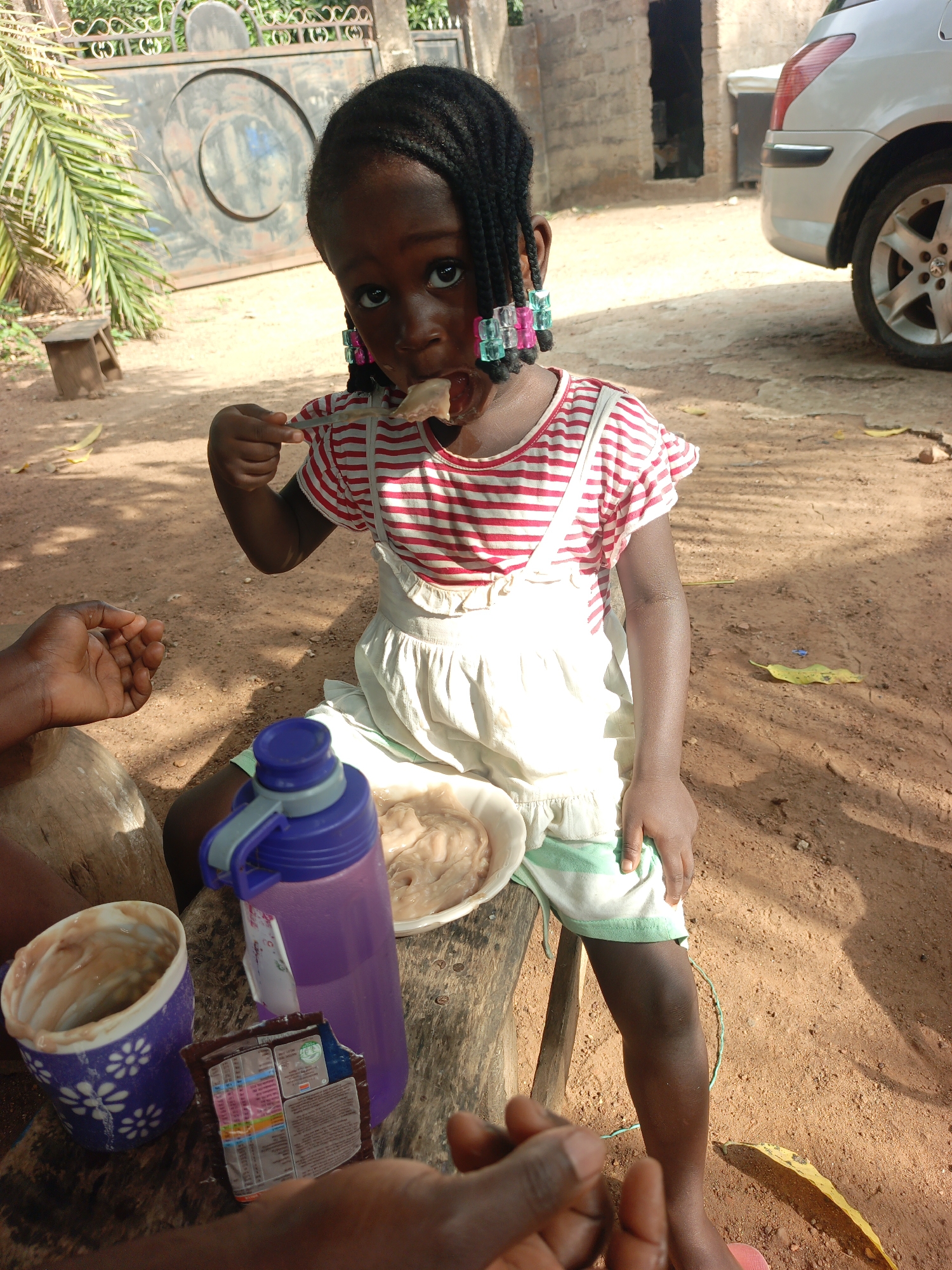 A young Black girl with beaded braids sits outdoors in Akwanga, Nigeria. She eats with a spoon. She wears a red and white striped shirt with a white dress and green-striped shorts. A blue water bottle and a cup containing food are next to her on the wooden seat. The ground is reddish-brown dirt. Behind her, a gate, a building, and a car are visible. An adult's hand is also visible in the foreground.