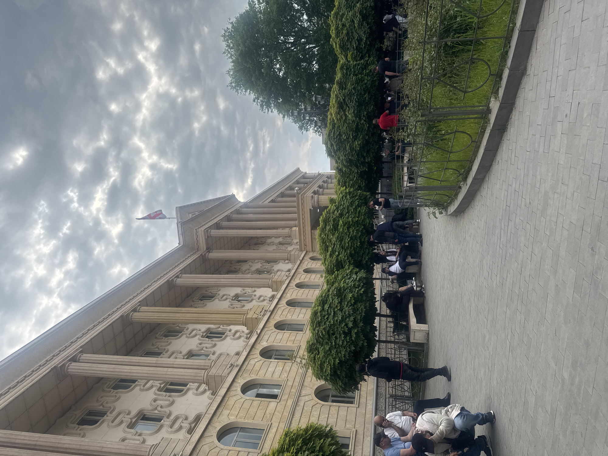 The image shows the Parliament of Georgia in Tbilisi. The building is a grand structure with classical architectural elements such as columns and arched windows. A Georgian flag is visible on the roof. A row of manicured trees lines the sidewalk next to the building. A metal fence runs along the edge of the sidewalk, separating it from a grassy area. Several people are present, some sitting on benches and others standing or kneeling on the pavement. Some people appear to be wearing masks. The sky is cloudy, indicating overcast weather.
