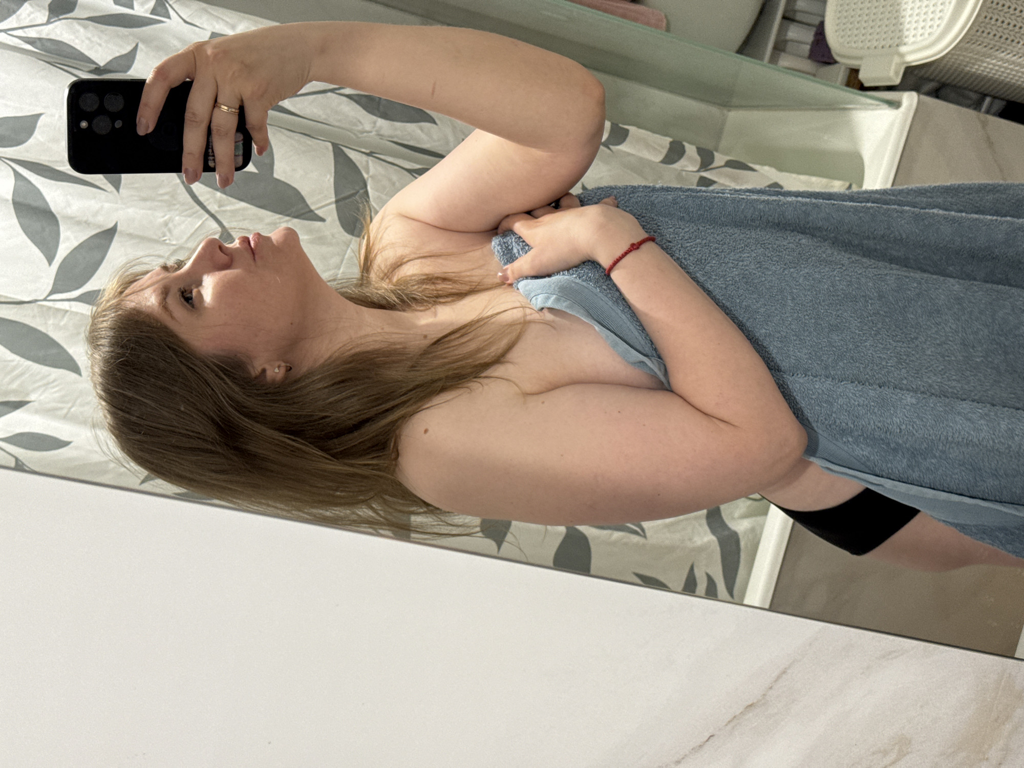 A woman is taking a selfie in a bathroom. She is holding a black smartphone in her right hand, which is positioned above her head and angled downward to capture her face and upper body. She is partially covered by a blue towel, and a red bracelet is visible on her wrist. The background includes a patterned shower curtain, bathroom fixtures, and a white countertop. The location is the city of Gornyi (Dvurechenskoe S/P), Russia.
