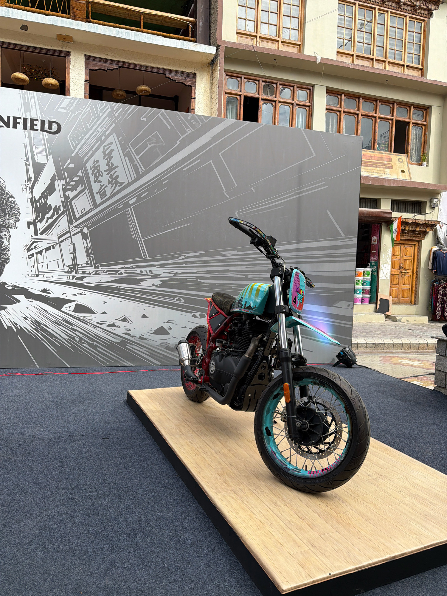 In the city of Leh, India, a custom Royal Enfield motorcycle is displayed on a wooden platform in front of a large graphic backdrop. The backdrop features a black and white illustration of a street scene with buildings and text. The motorcycle itself is vibrantly colored with teal, yellow, and pink accents, and has a custom paint job. It is a scrambler-style bike with knobby tires. In the background, a traditional Ladakhi building with wooden windows and balconies is visible. The street in front of the building is paved with cobblestones, and there are shops with merchandise displayed outside. The overall scene appears to be an outdoor exhibition or event.