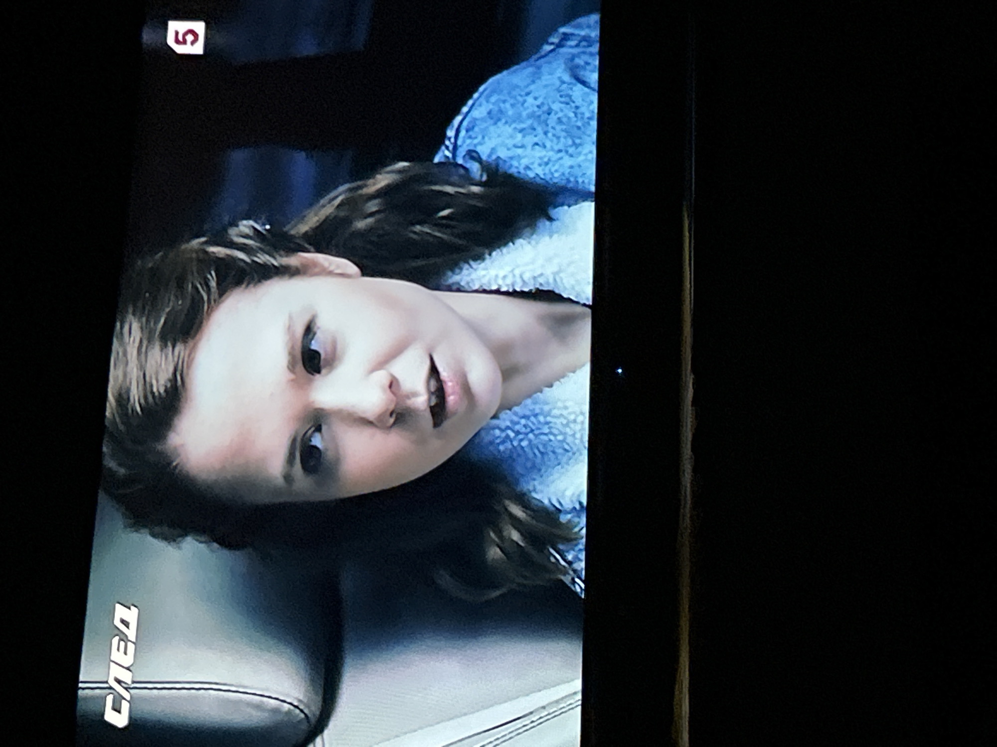 The image shows a television screen displaying a scene with a young woman. She has light brown hair pulled back, with some strands framing her face. She is wearing a light blue denim jacket with a white fuzzy collar. The background appears to be the interior of a car, with a dark headrest visible on the left side of the screen. In the upper left corner of the screen, a logo that reads "СЛЕД" is visible. In the upper right corner, a small white rectangle with a red number "5" inside is displayed. The overall lighting suggests an indoor or nighttime setting. There is no discernible information about the city of Gelendzhik, Russia, within the image itself.