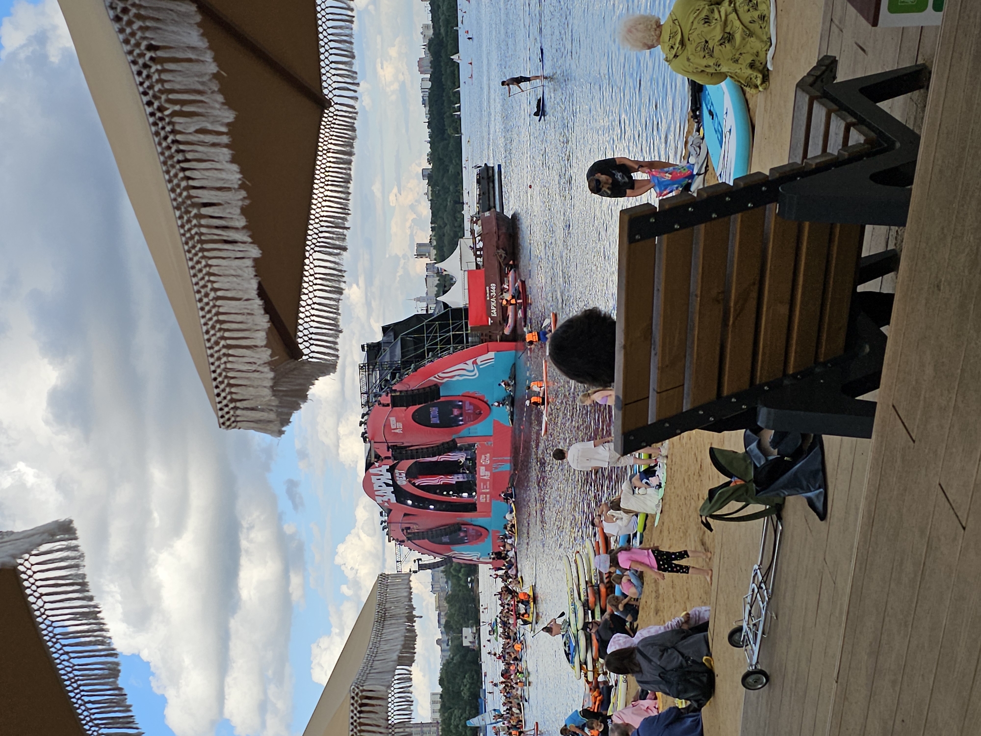 The image depicts a lively scene at a waterfront event in Khimki, Russia. In the background, a large, red, arch-shaped stage with the Cyrillic text "ЗАРЯДКА" (Zaryadka) and "ФЕСТ" (Fest) is set up on the water, suggesting a festival or concert. Numerous people are engaged in water activities, including paddleboarding and kayaking, on a wide body of water. The foreground shows a sandy beach with people relaxing, sitting on towels, and a few paddleboards are stacked on the sand. A wooden deck in the immediate foreground features empty seating, with a wheeled trolley and bags placed to the side. The sky is partly cloudy, with a mix of blue sky and white clouds. The overall atmosphere appears to be that of a summer day at a popular outdoor venue.